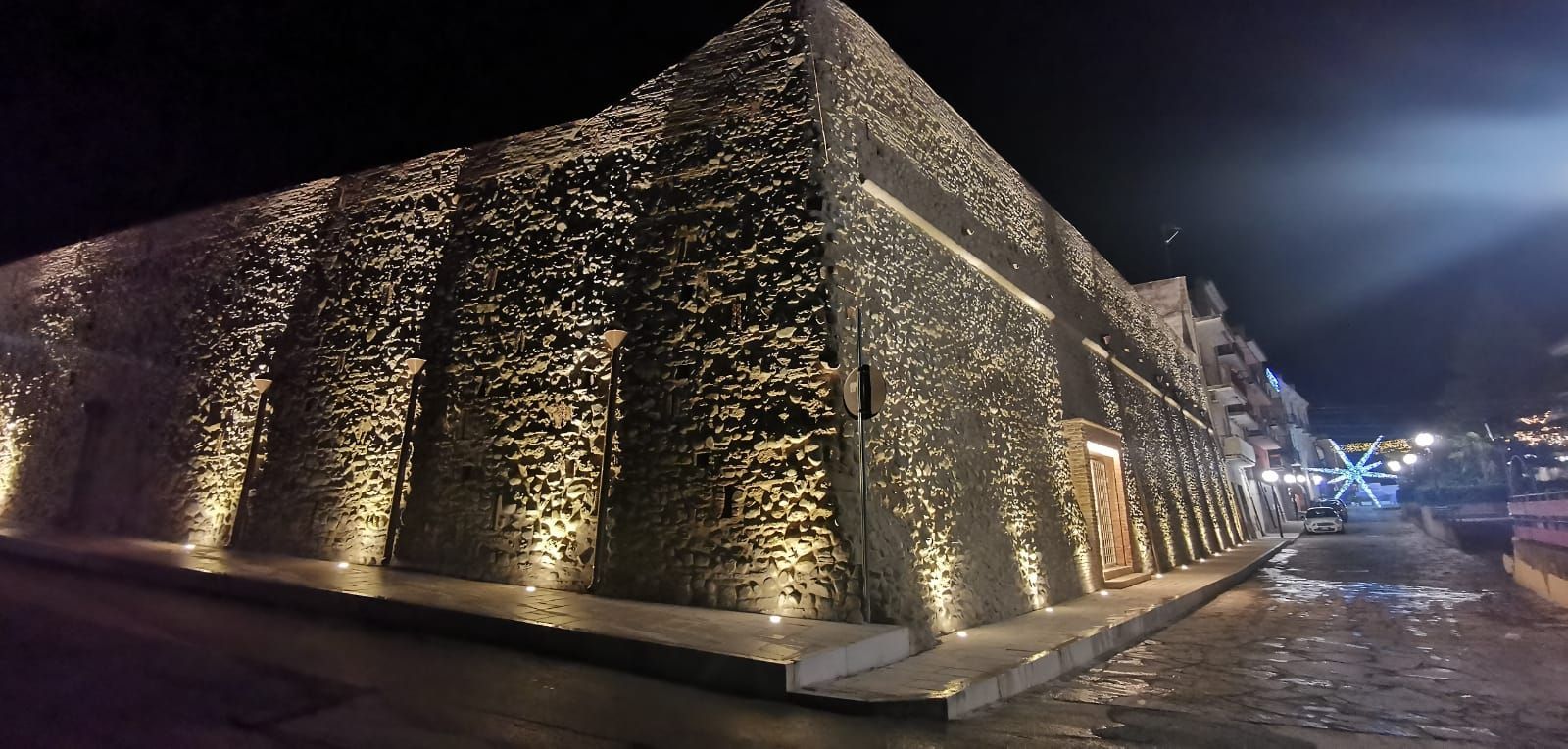 Vista notturna di un edificio in pietra con faretti che ne illuminano la facciata strutturata. Sono visibili la strada e l'acqua.