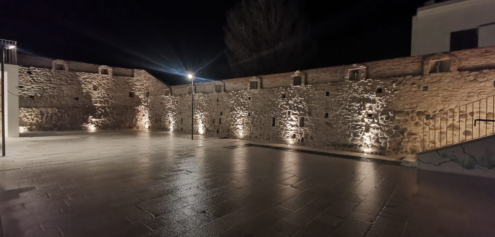 Muro di pietra illuminato di notte dai lampioni che si riflettono sul marciapiede bagnato.