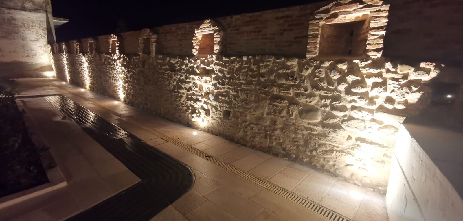 Vista notturna di un muro di pietra con nicchie illuminate e sentiero.