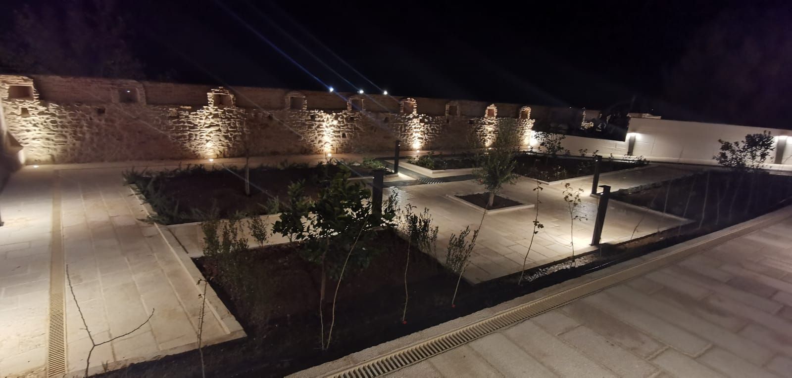 Vista notturna di un cortile con muri in pietra illuminati e paesaggio circostante, e faretti in primo piano.
