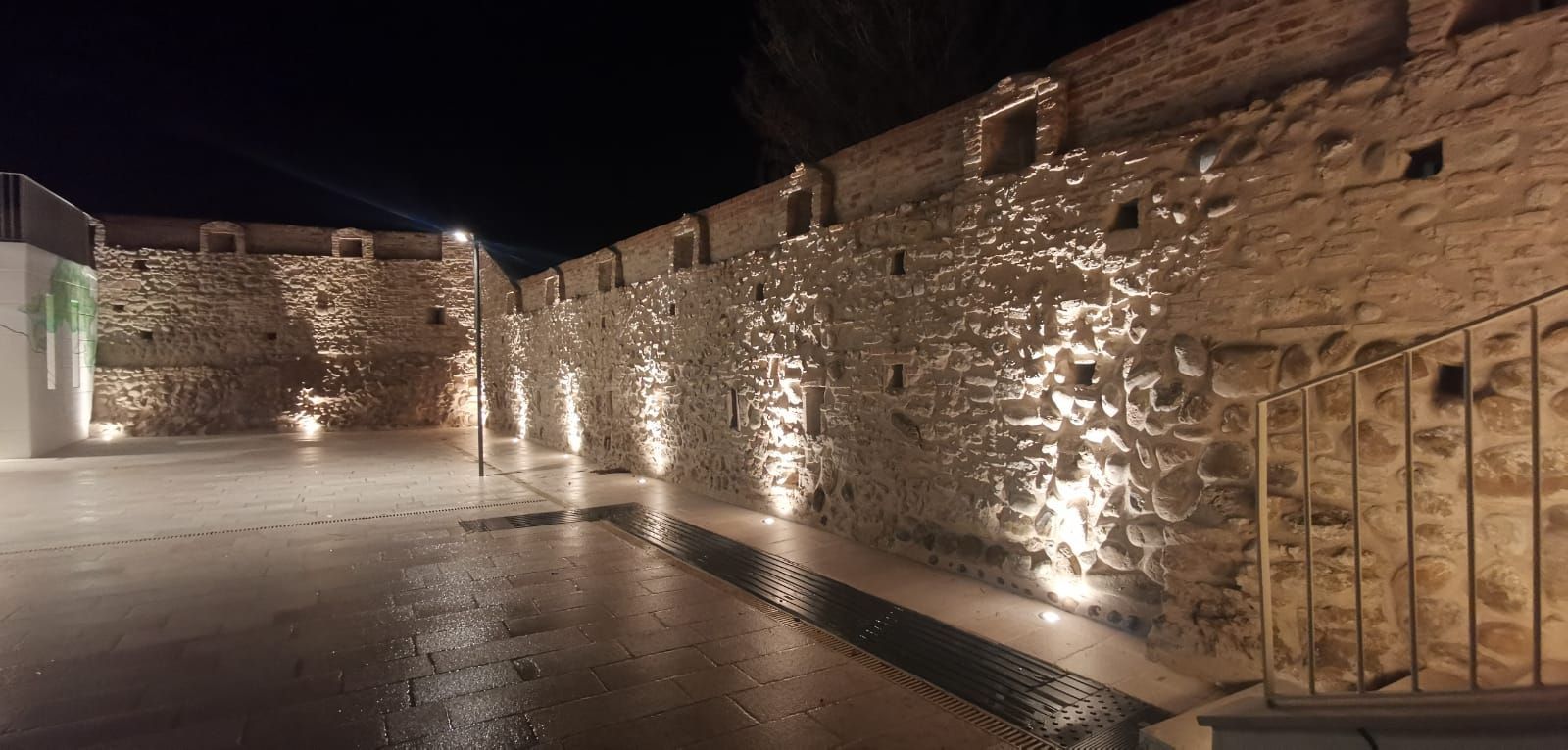Vista notturna di un muro di pietra illuminato da faretti, con una piazza umida di fronte.