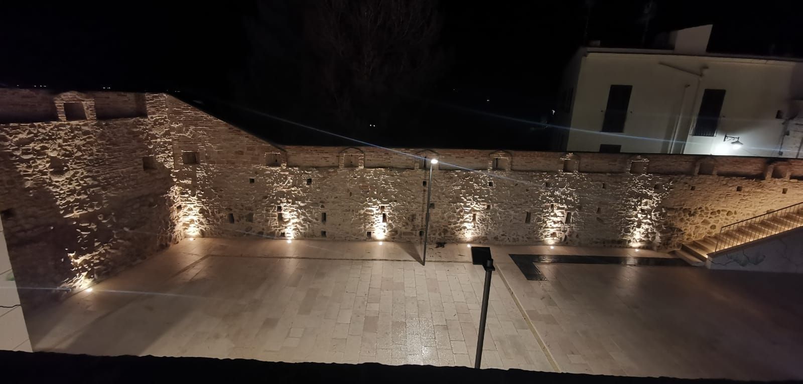 Ripresa notturna di un muro di pietra illuminato da faretti. Di fronte al muro si trova un cortile e sulla destra un edificio.