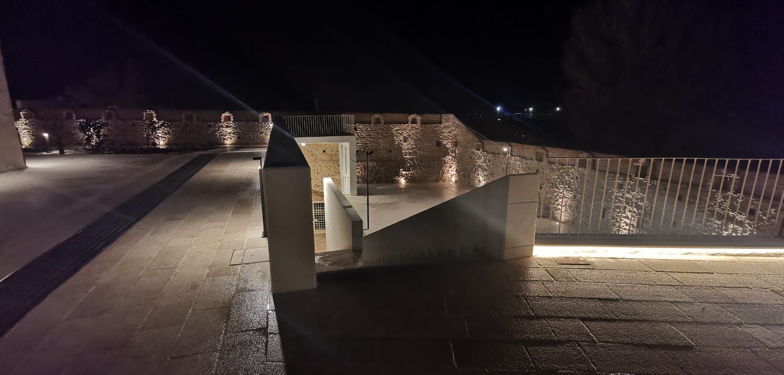 Vista notturna di un esterno moderno con pareti in pietra illuminate, una scala e una superficie bagnata e riflettente.