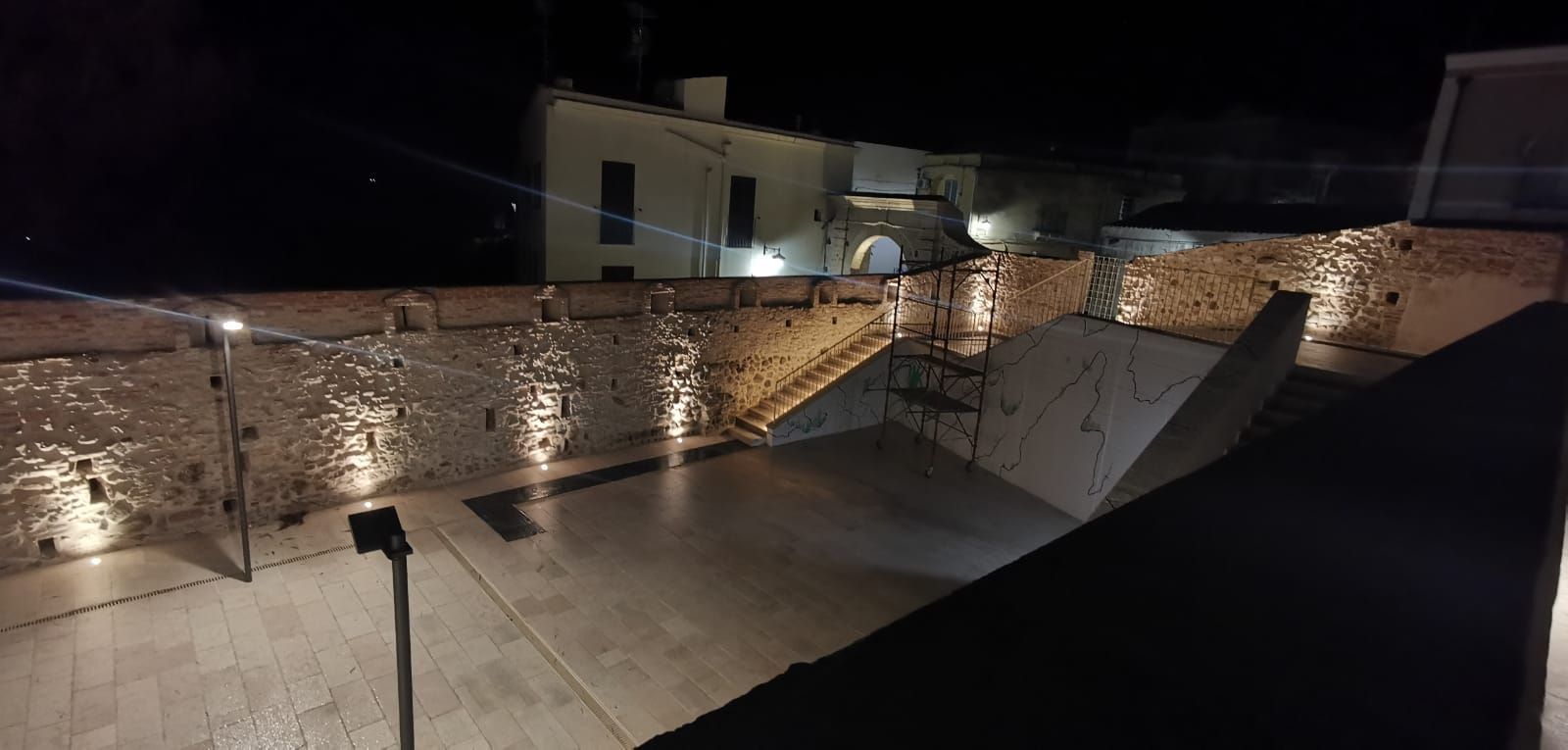Ripresa notturna di una terrazza in pietra con illuminazione a parete e un edificio sullo sfondo.