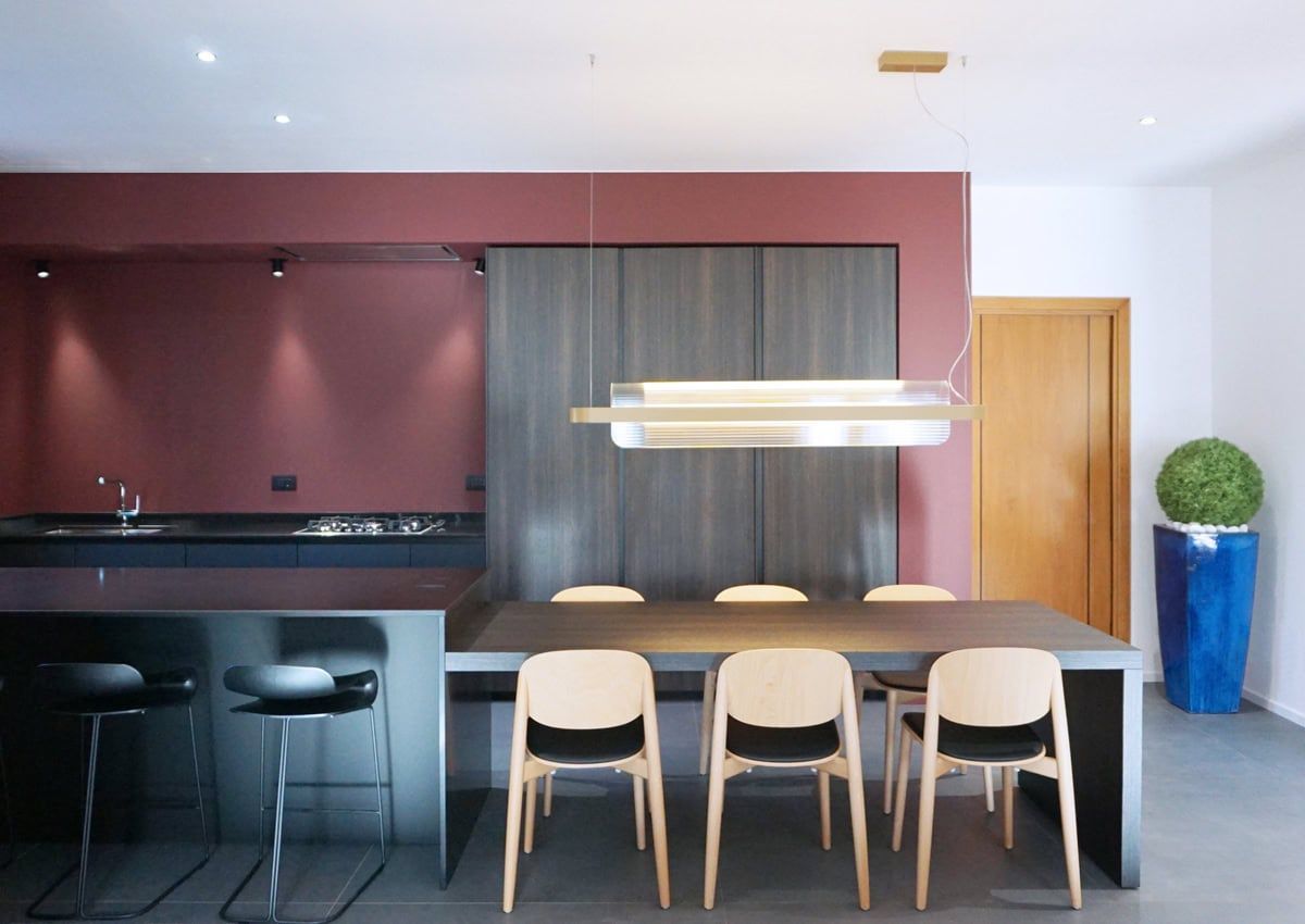 Cucina e sala da pranzo moderne con parete bordeaux, mobili scuri e sedie neutre.