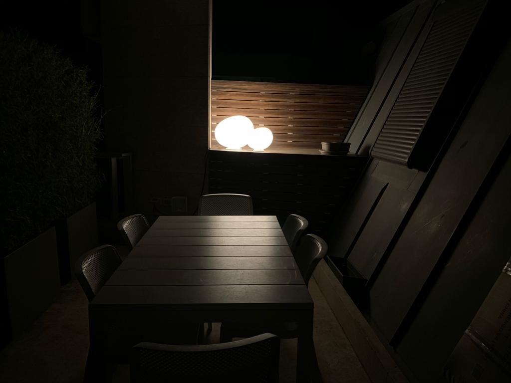 Zona pranzo esterna buia con un lungo tavolo e sedie, illuminata da luci sferiche luminose.