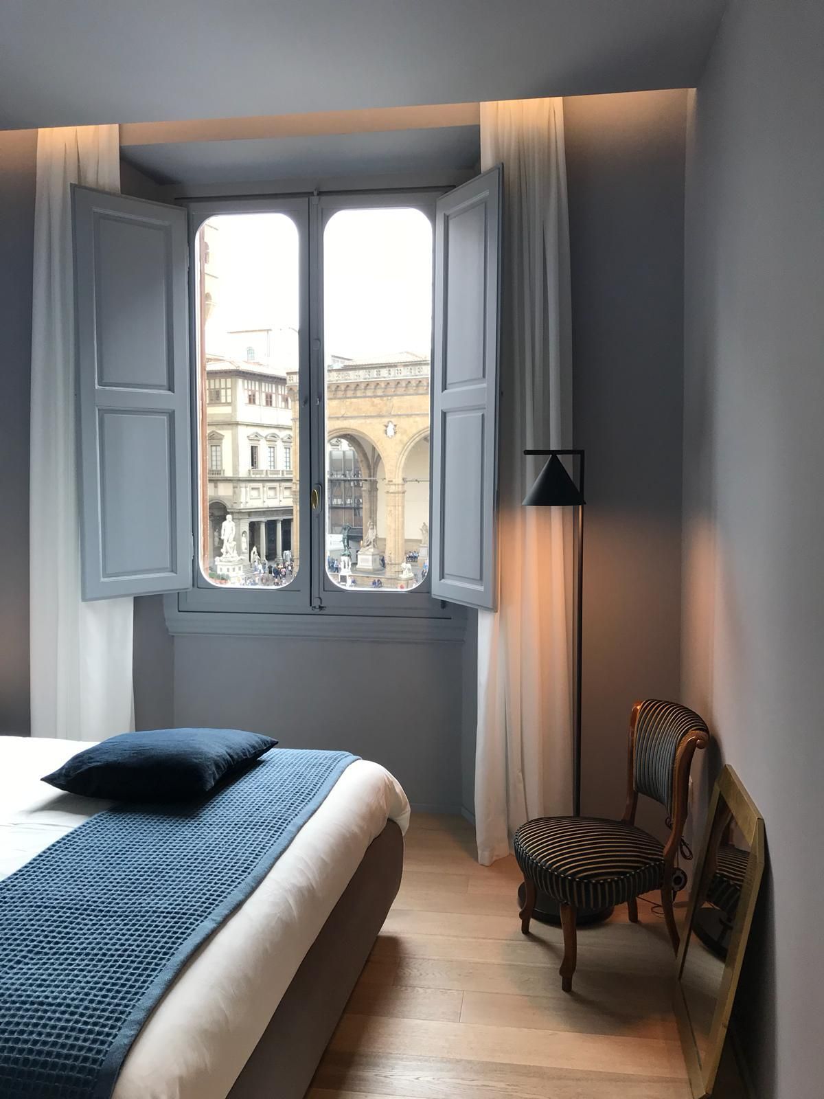 Camera da letto con finestra aperta che si affaccia su una strada europea. Persiane, tende, letto e sedia azzurri.