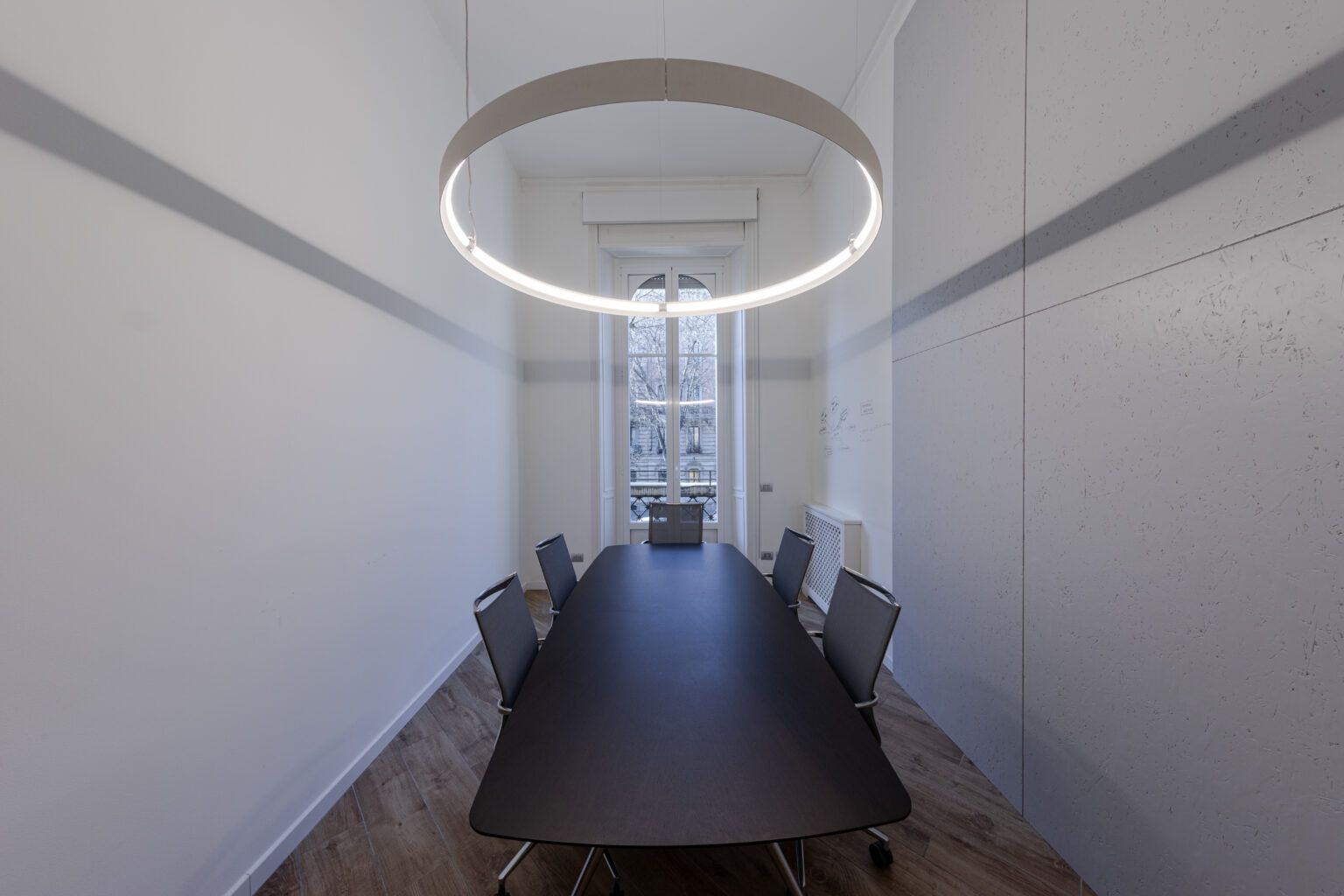 Sala conferenze con lungo tavolo nero, sedie e lampada circolare sopraelevata. Finestra in fondo.