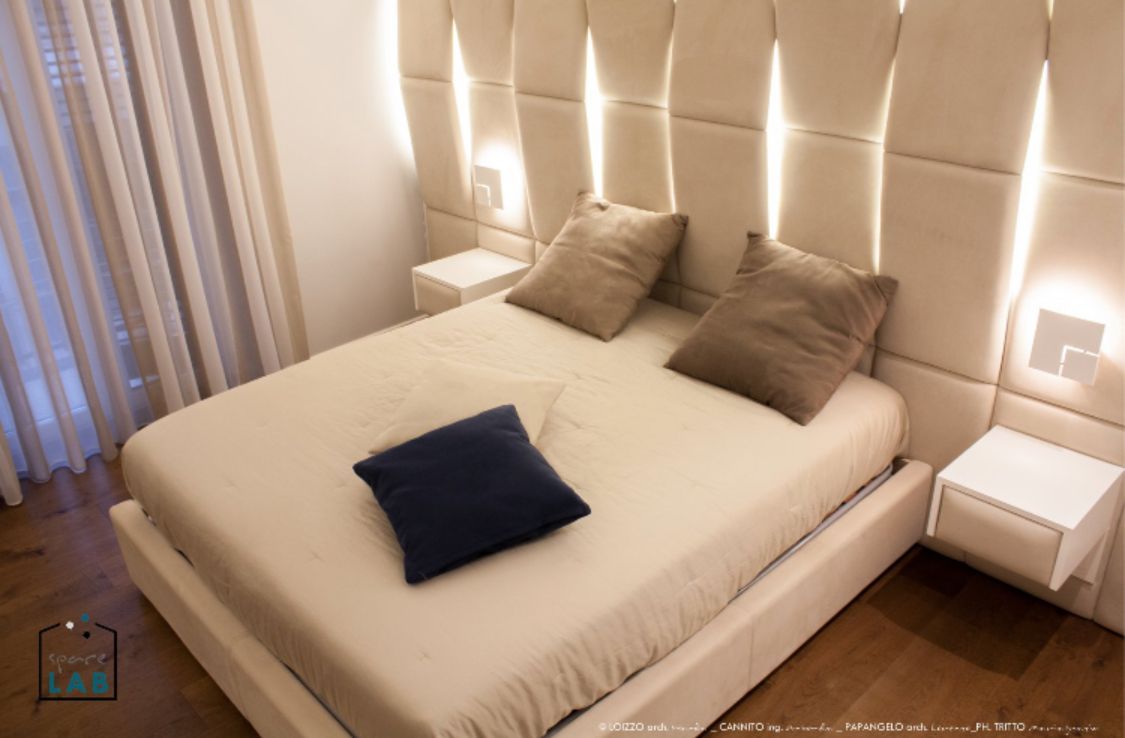 Camera da letto con letto bianco, testiera beige con luci, due comodini e un cuscino blu scuro.
