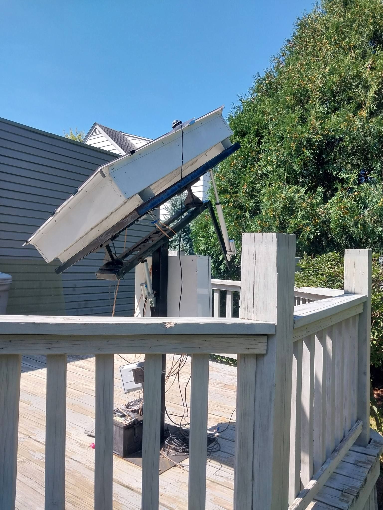 Solar panel mounted on a deck, angled towards the sun, with wooden railing. Blue sky, green trees in background.