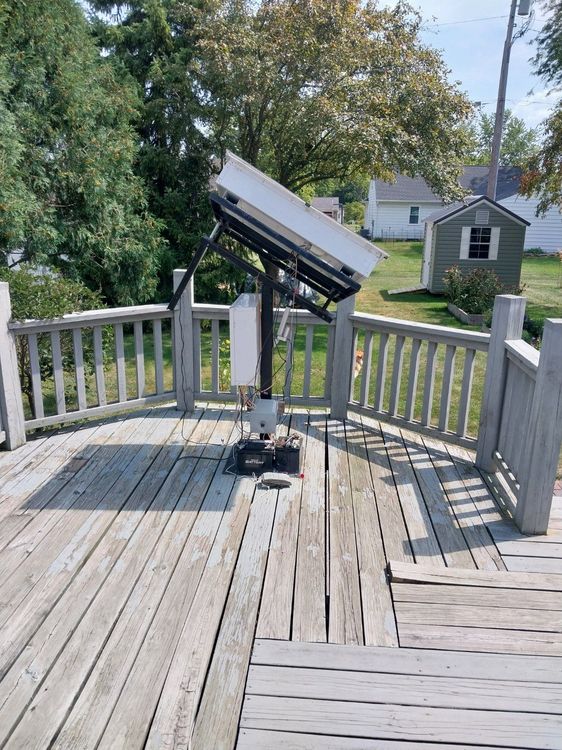 Solar panel on a deck; tilted towards the sky, on a gray, weathered deck. Trees and houses in the background.