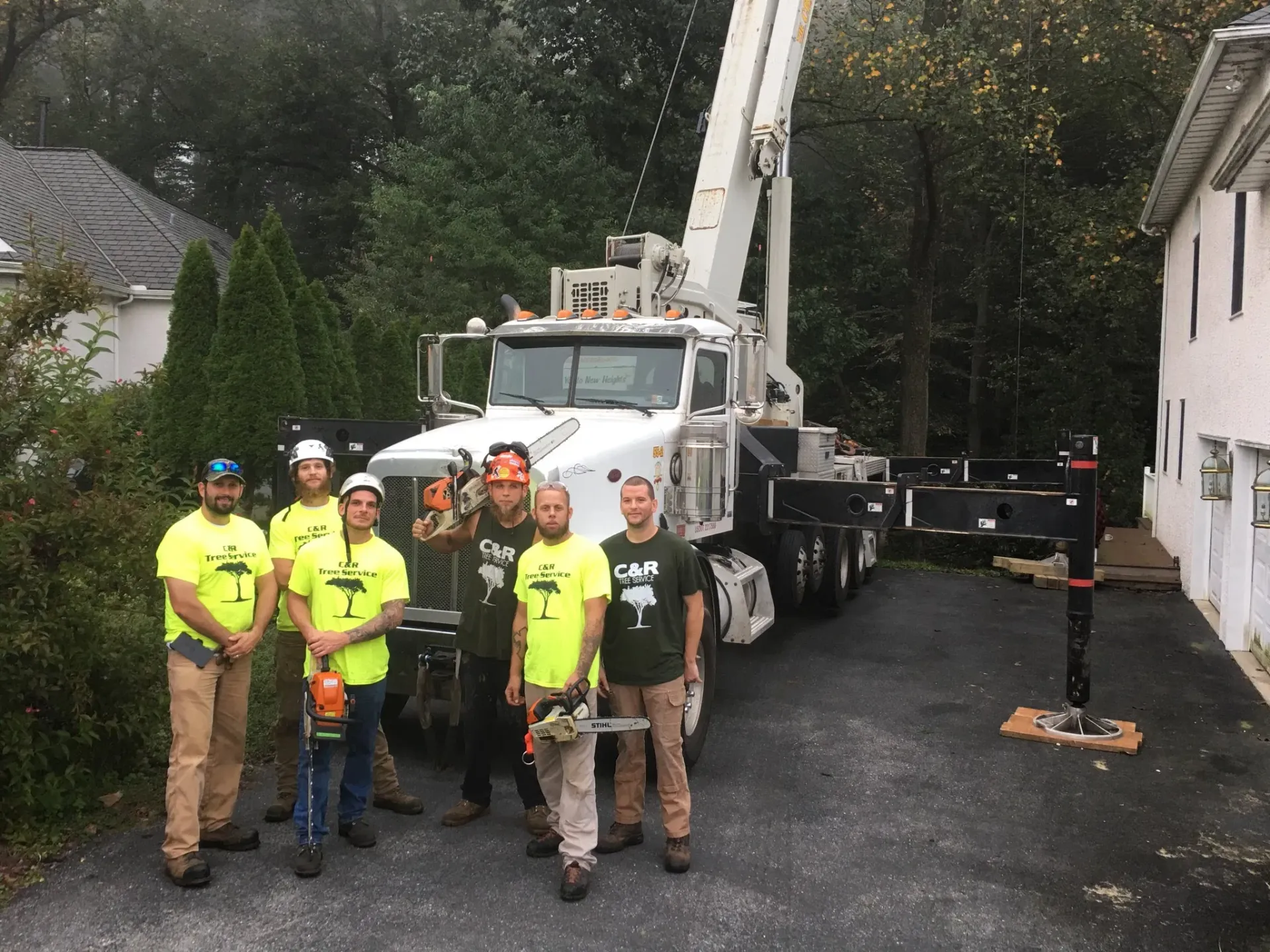 Tree service crew stands in front of a truck-mounted crane. Crew members hold chainsaws in a residential setting.