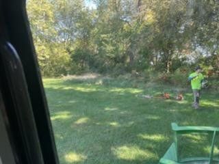 Person in neon shirt using a tool on a grassy field near trees.