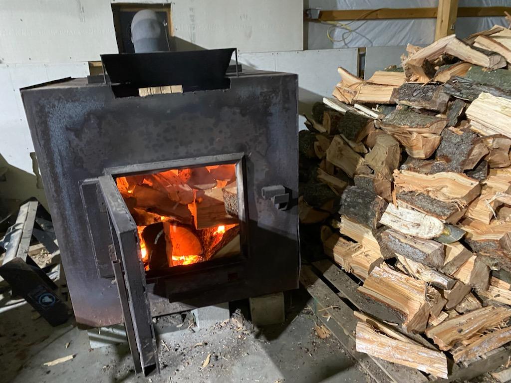 Wood-burning stove with open door, flames visible, next to a stack of firewood in a building.