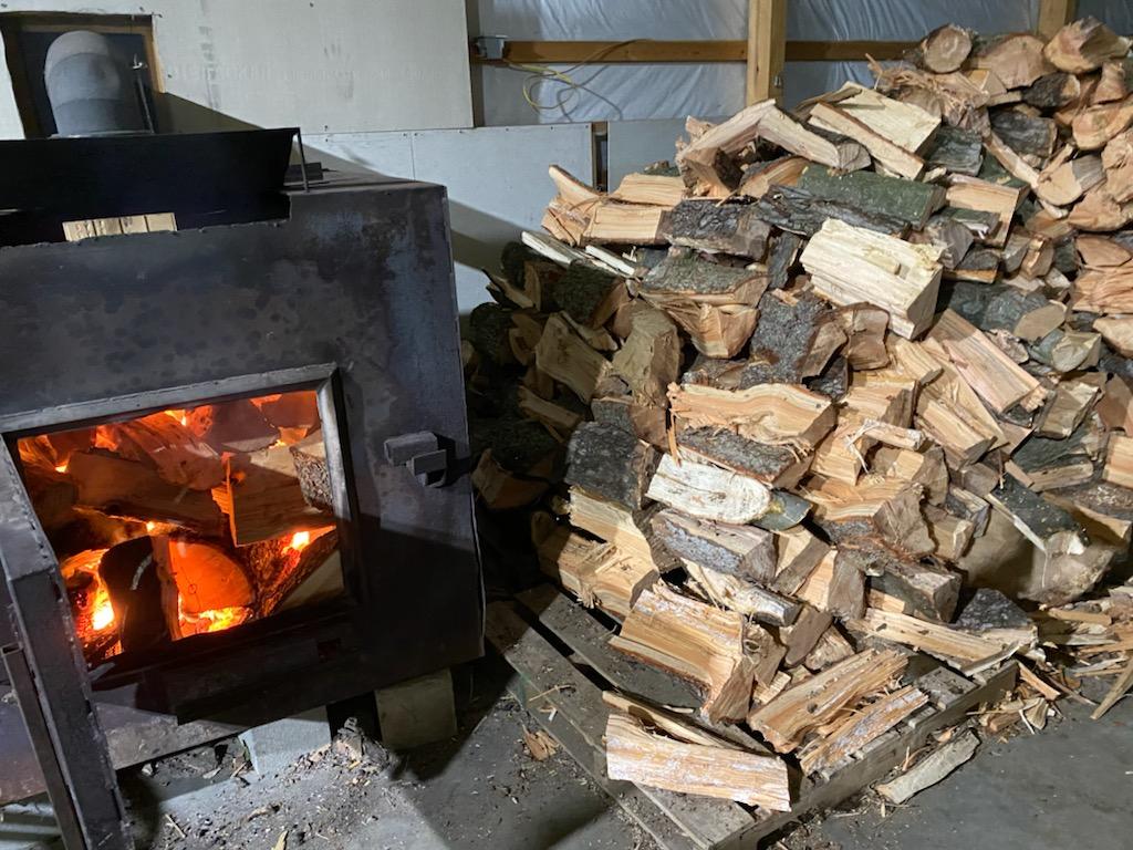 A wood-burning stove with open door, burning logs, beside a large pile of firewood in a wooden structure.