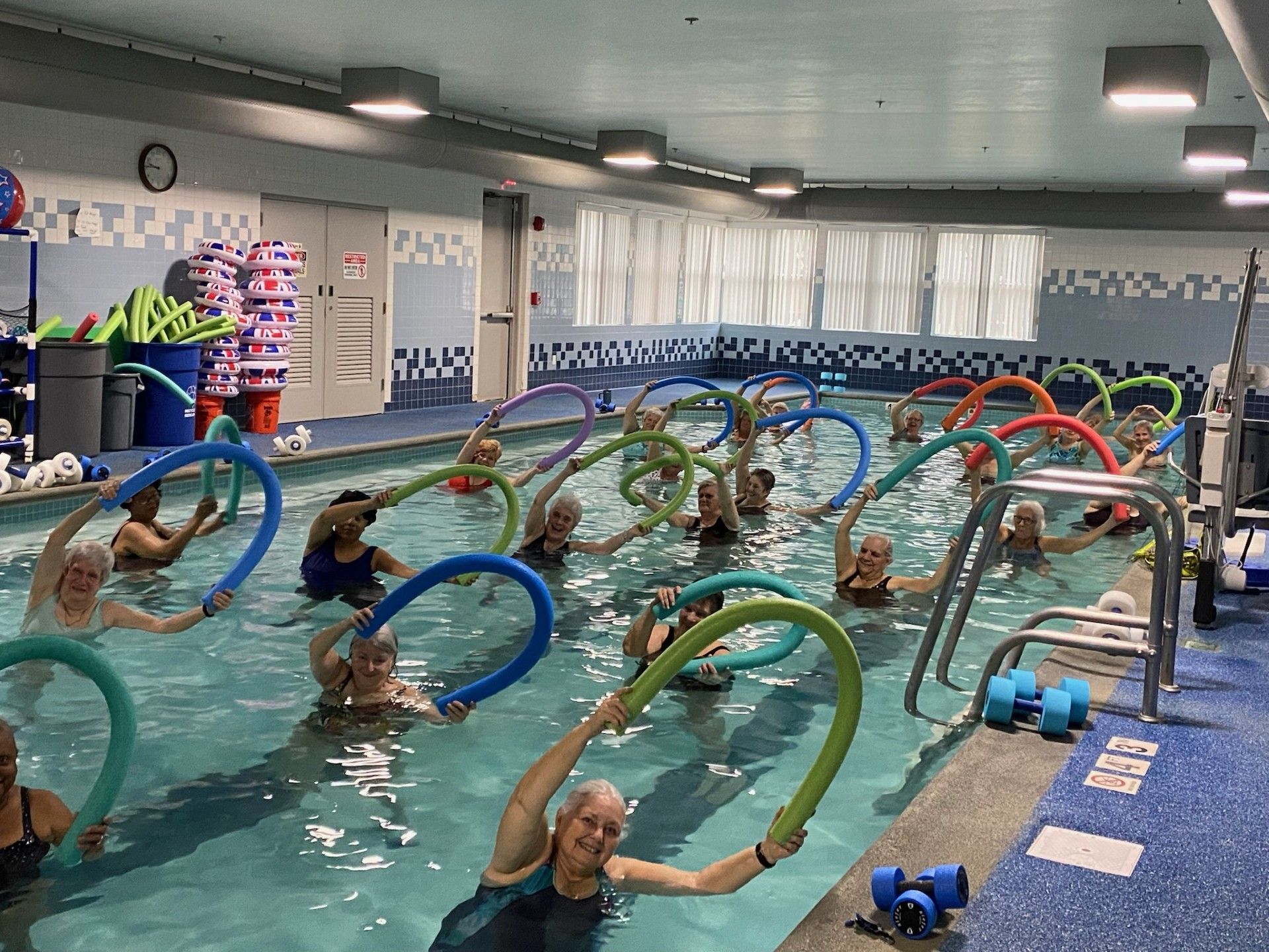 A group of people in an indoor pool performing water aerobics exercises with green foam pool noodles.