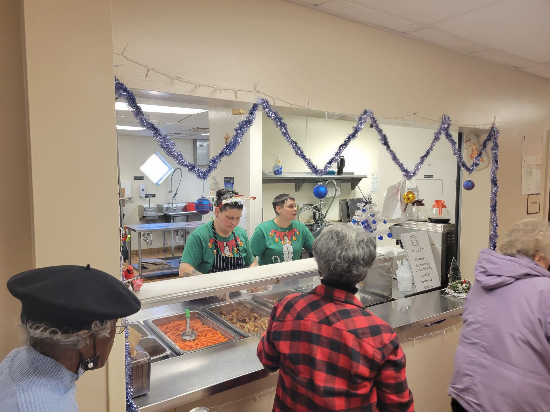 People wait in a cafeteria line as staff serve food behind a counter decorated with blue tinsel and holiday ornaments.