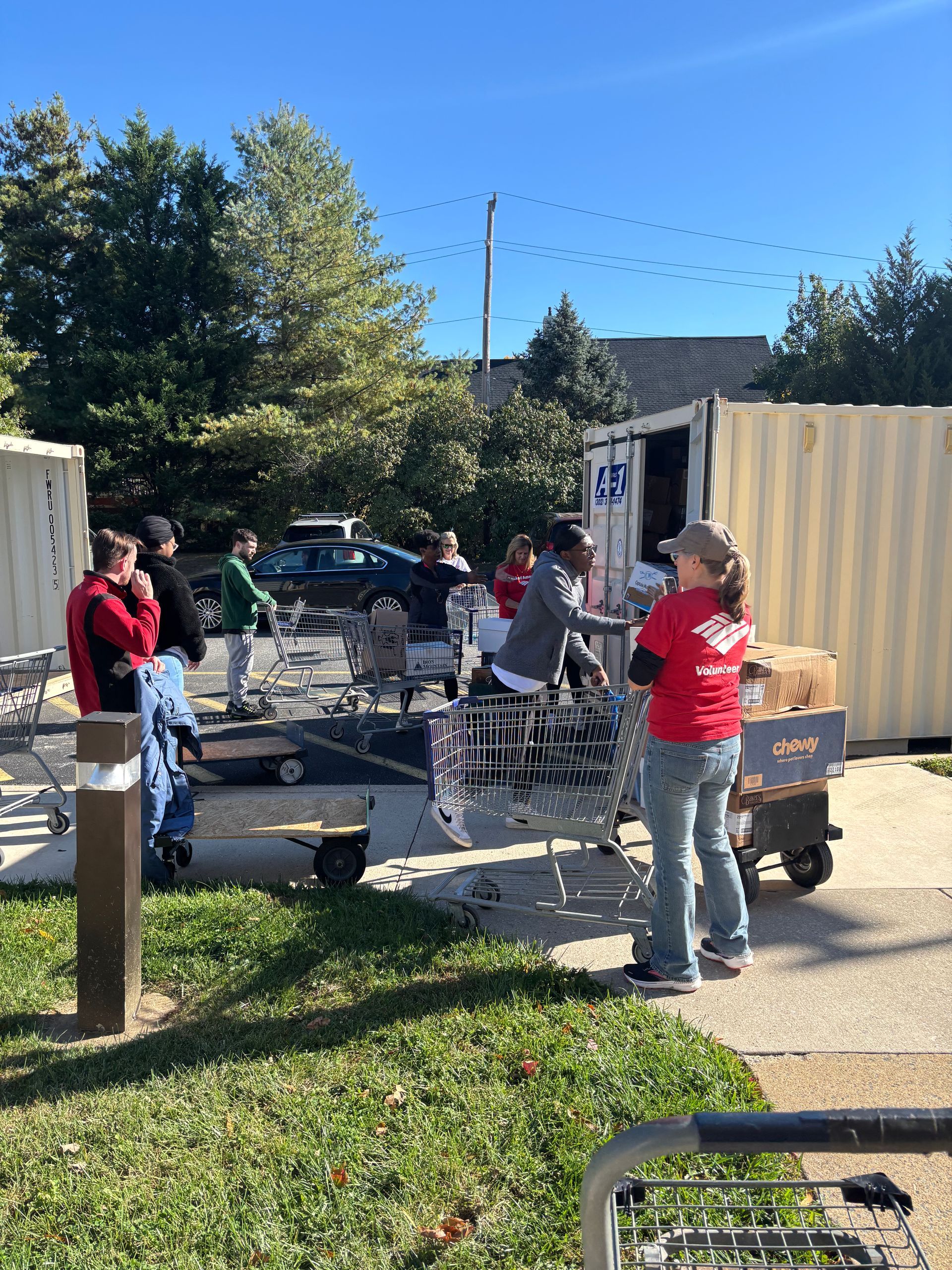 Volunteers in red shirts organize food donations from a storage container into carts on a sunny day.