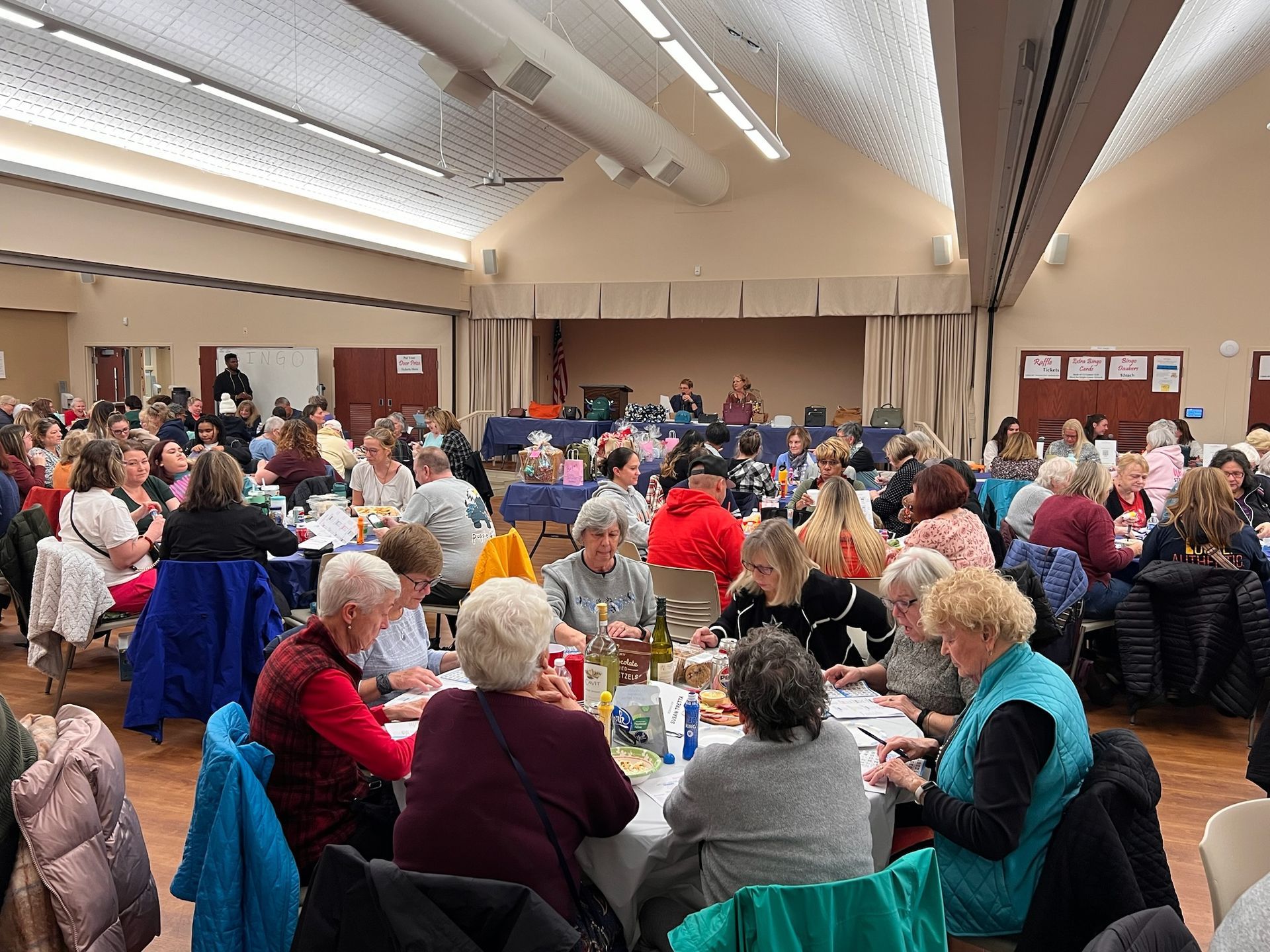 A large group of people seated at round tables in a brightly lit community hall, enjoying a meal and socializing.