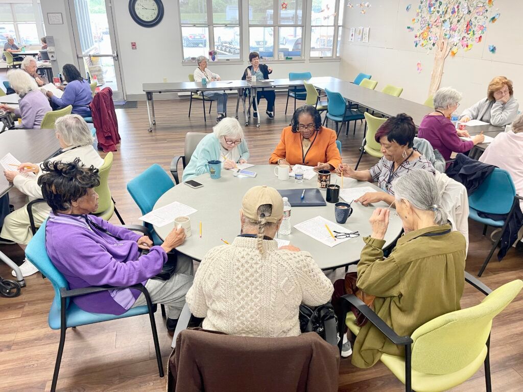 A group of people sit at a large round table in a brightly lit community room, reading and talking during a gathering.