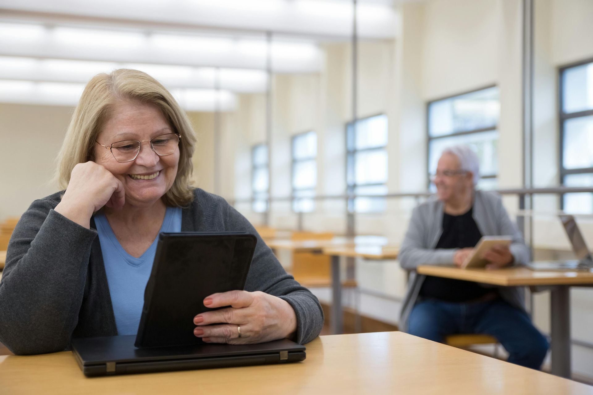 A person smiling while reading from a digital tablet in a bright library setting, with another person working in the back.