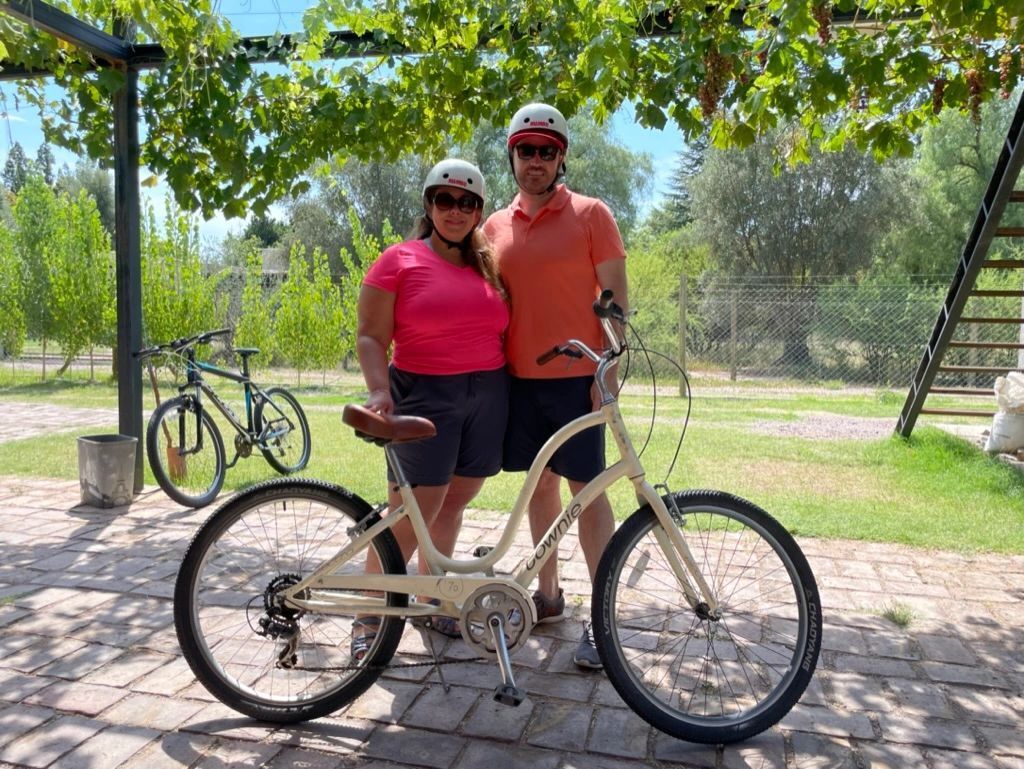 Two people standing beside a cream-colored bicycle in a sunny park, with trees and a dirt path behind them.