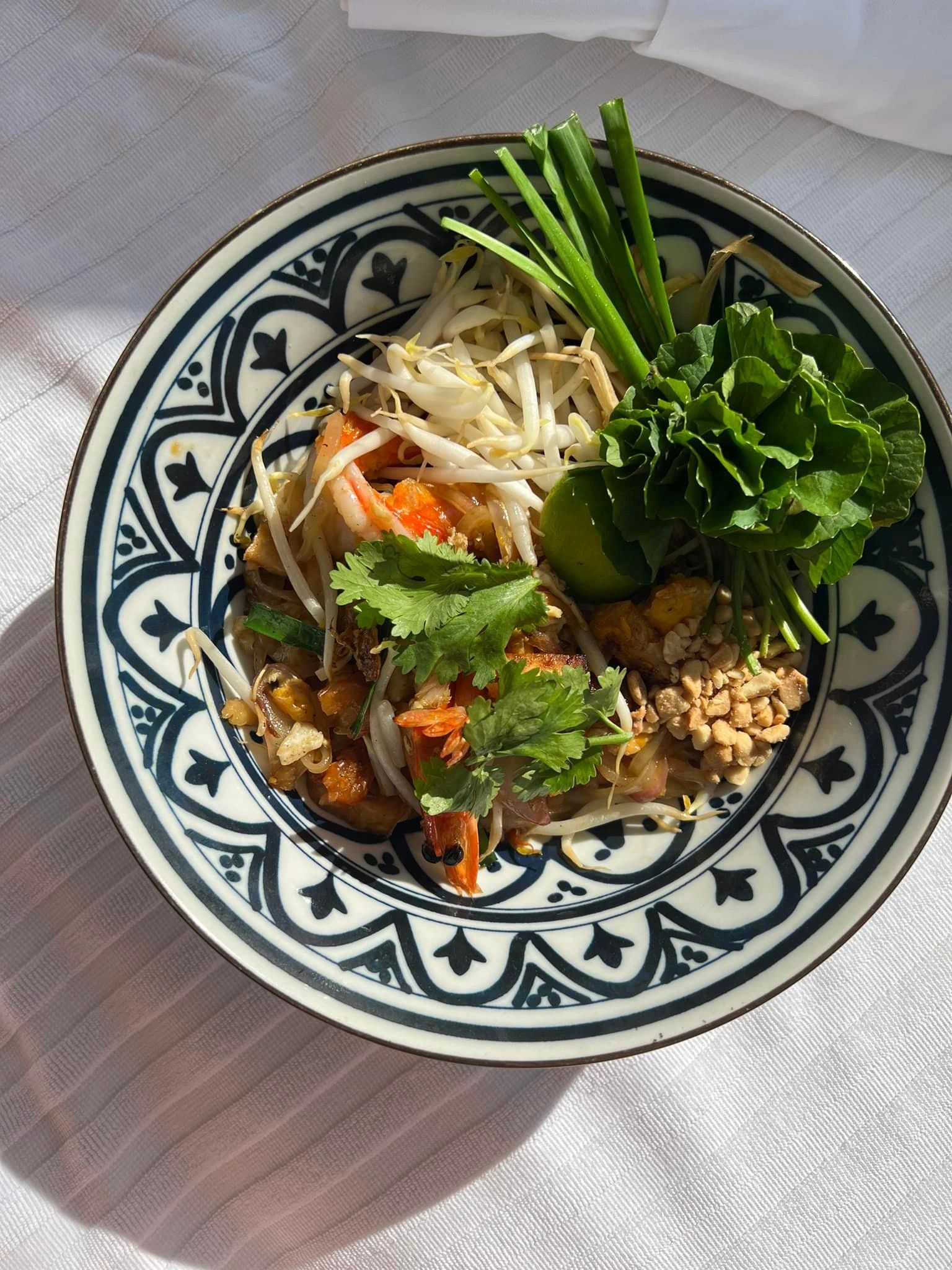 Decorative bowl of fresh noodles with herbs, bean sprouts, lime, and peanuts on a sunlit table