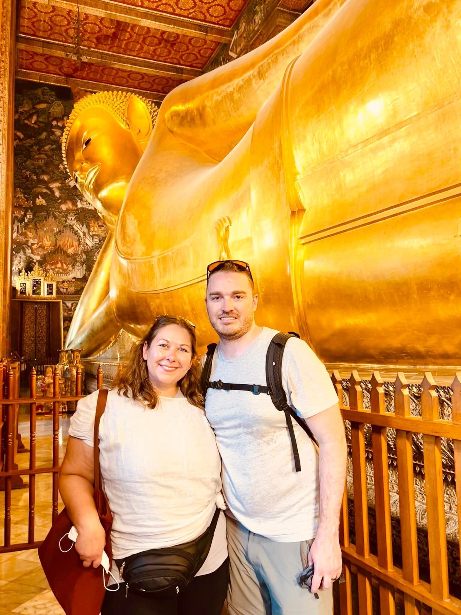 Two people posing beside a large golden Buddha statue inside a temple, smiling.