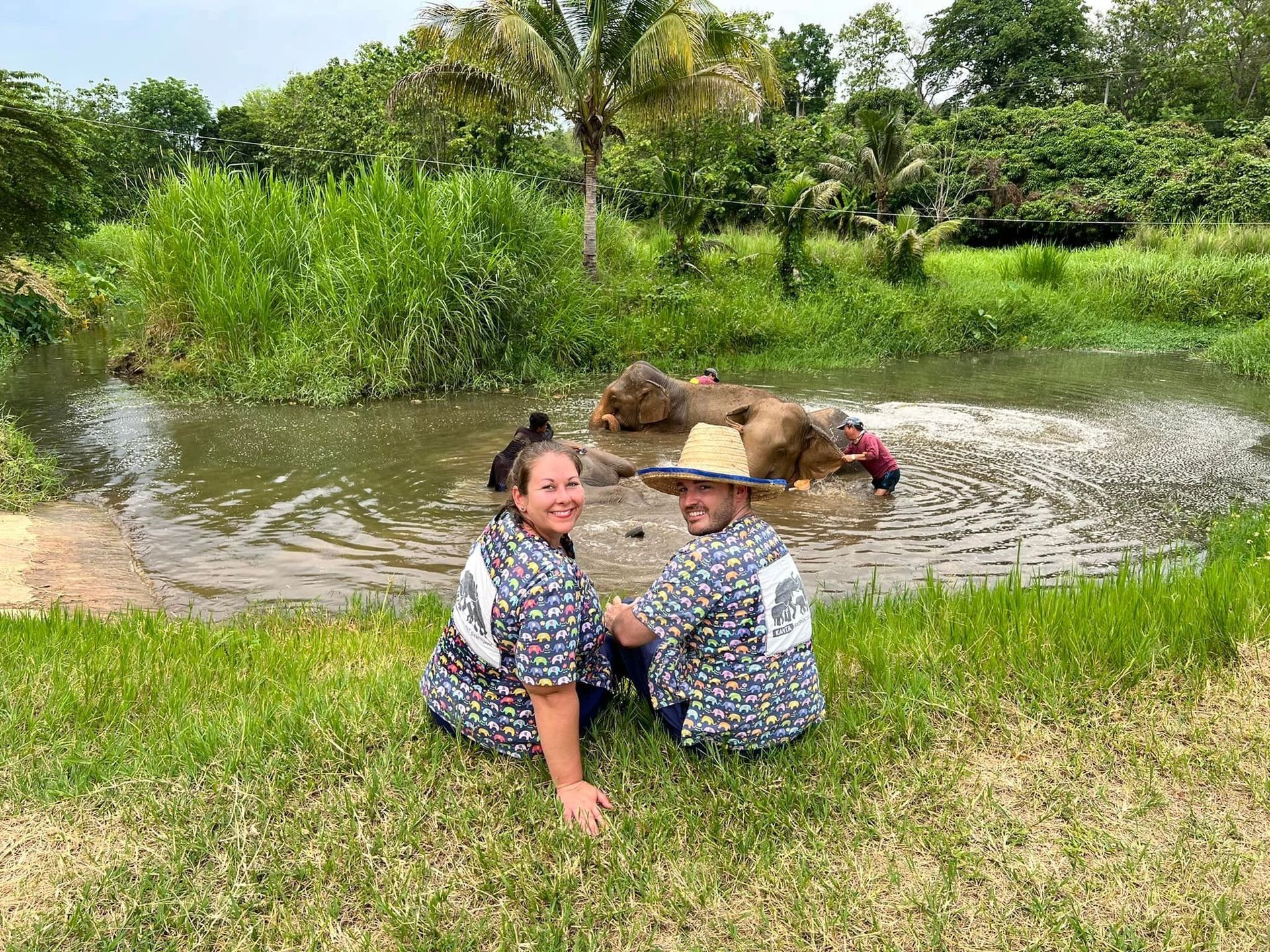 Two people sitting by a pond, smiling, with a water buffalo in the background.