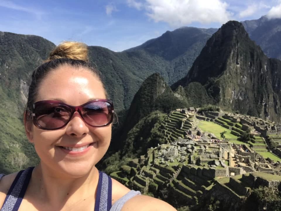 Smiling woman in sunglasses overlooking Machu Picchu ruins under a blue sky