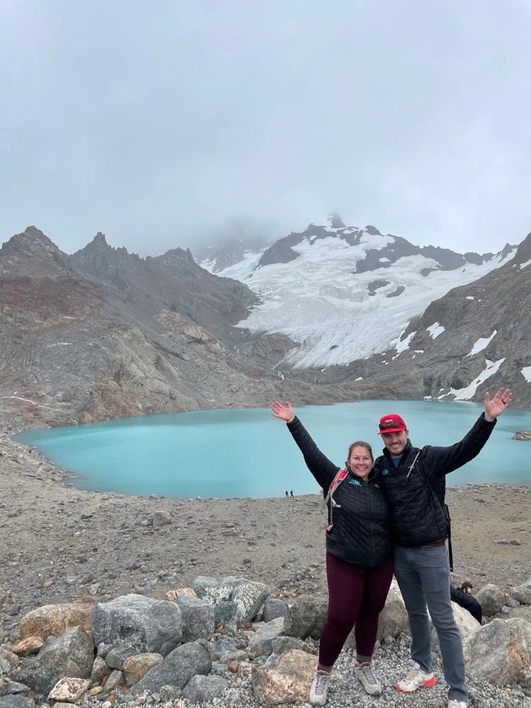 Two hikers pose with arms raised beside a turquoise glacial lake and snowy mountains.