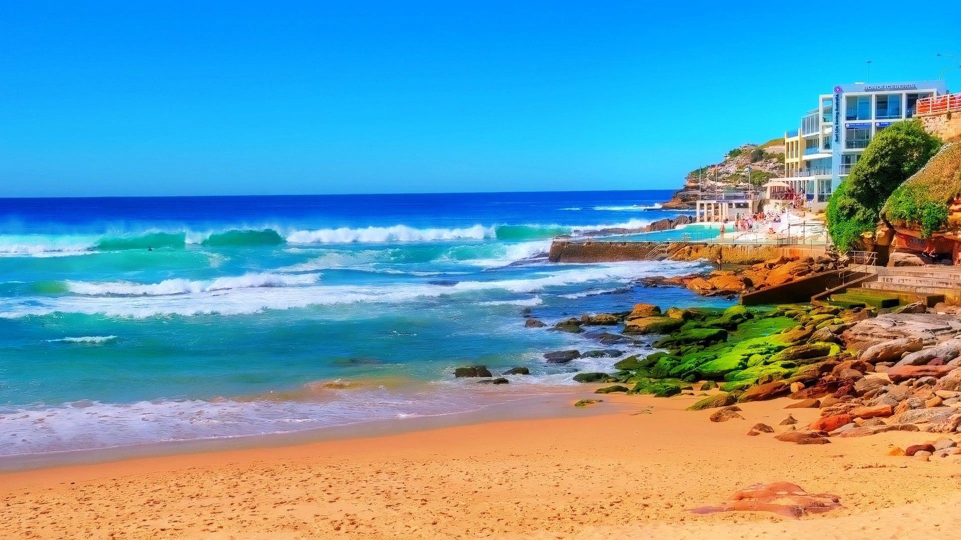 Sunny beach with turquoise waves, sandy shore, rocky coastline, and buildings along the right side.