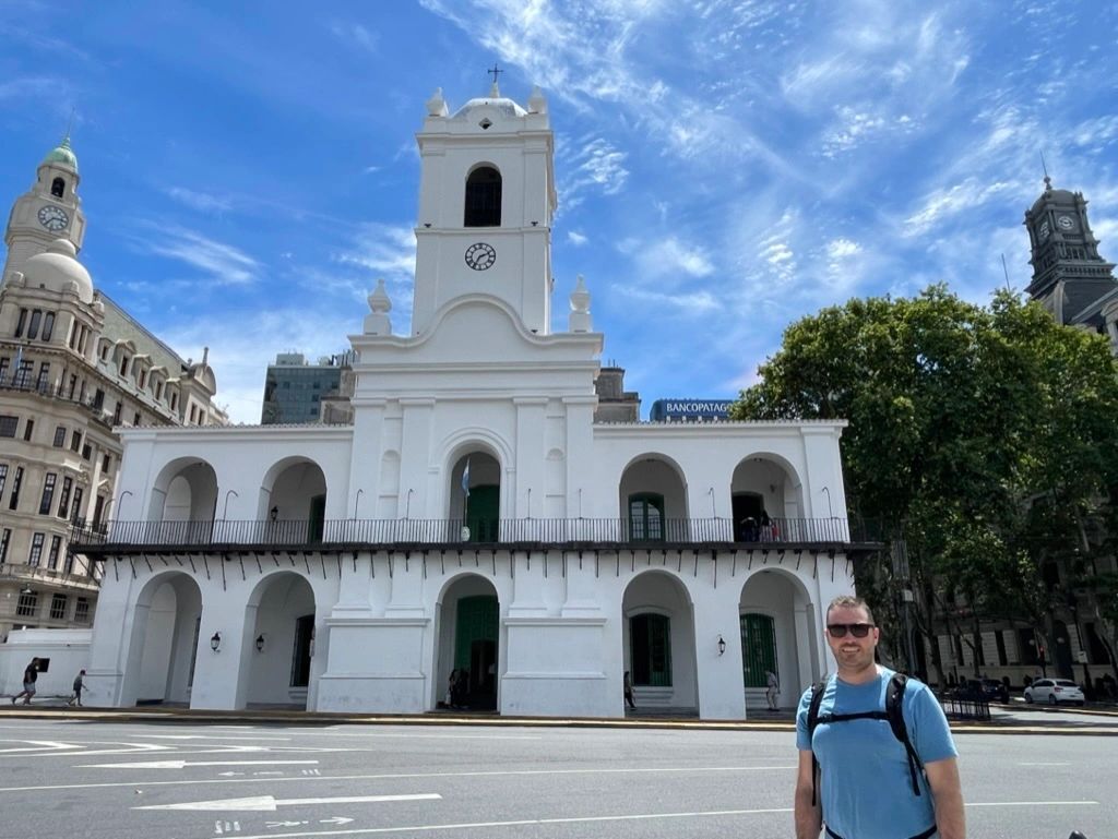 White historic building with a clock tower in a sunny city square; man in blue shirt stands in front.