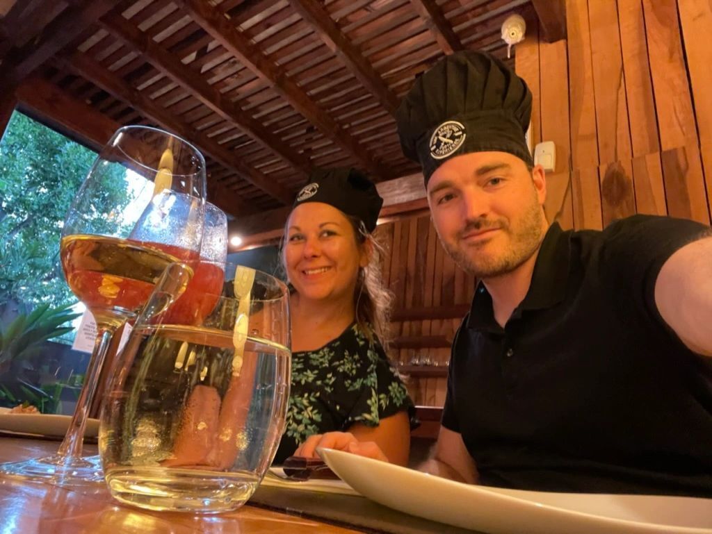 Two people in chef hats smiling at a wooden table with a large glass drink jug and a plate.