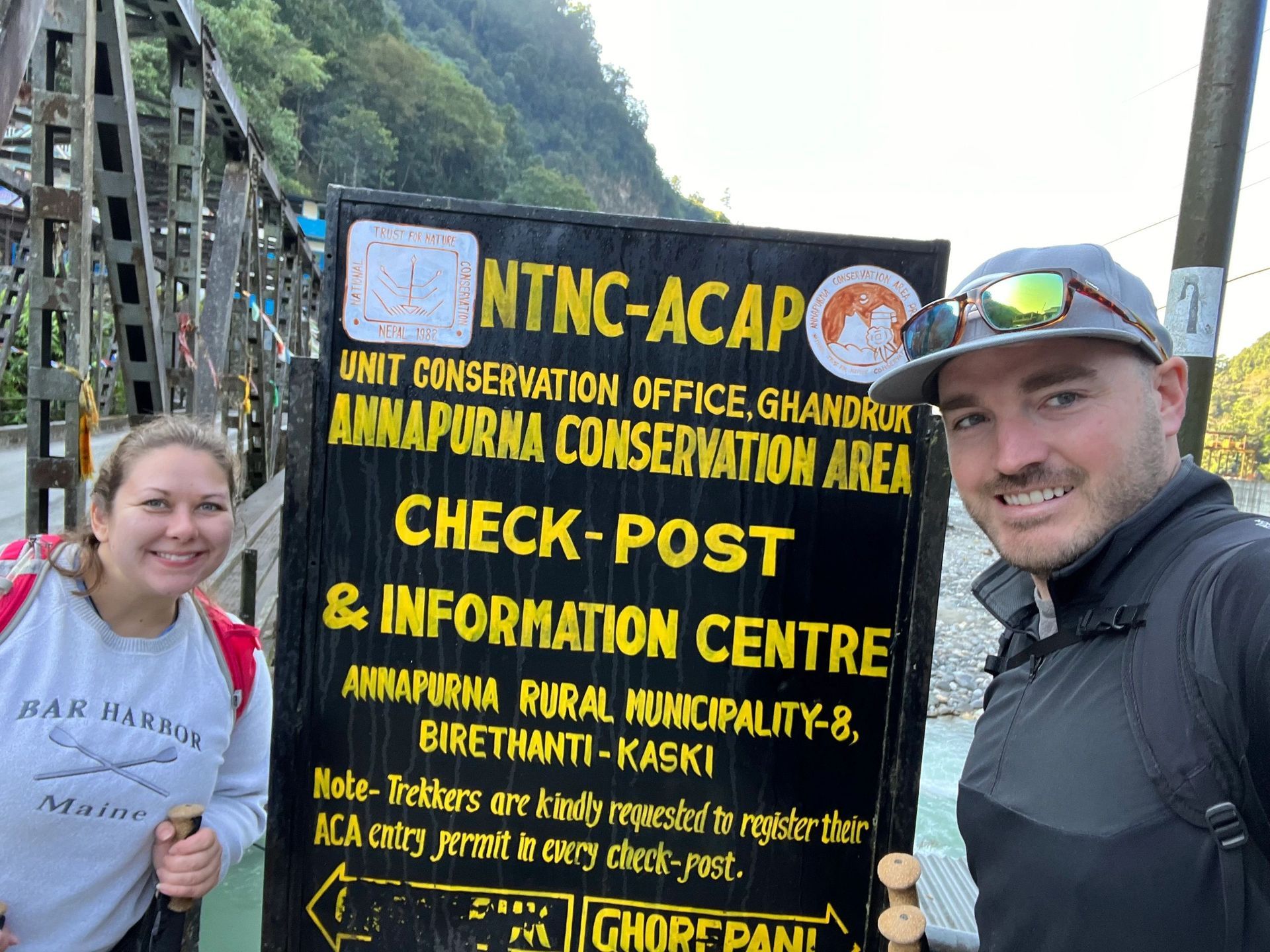 Two hikers posing beside a black-and-yellow Annapurna Conservation Area sign at a trailhead.