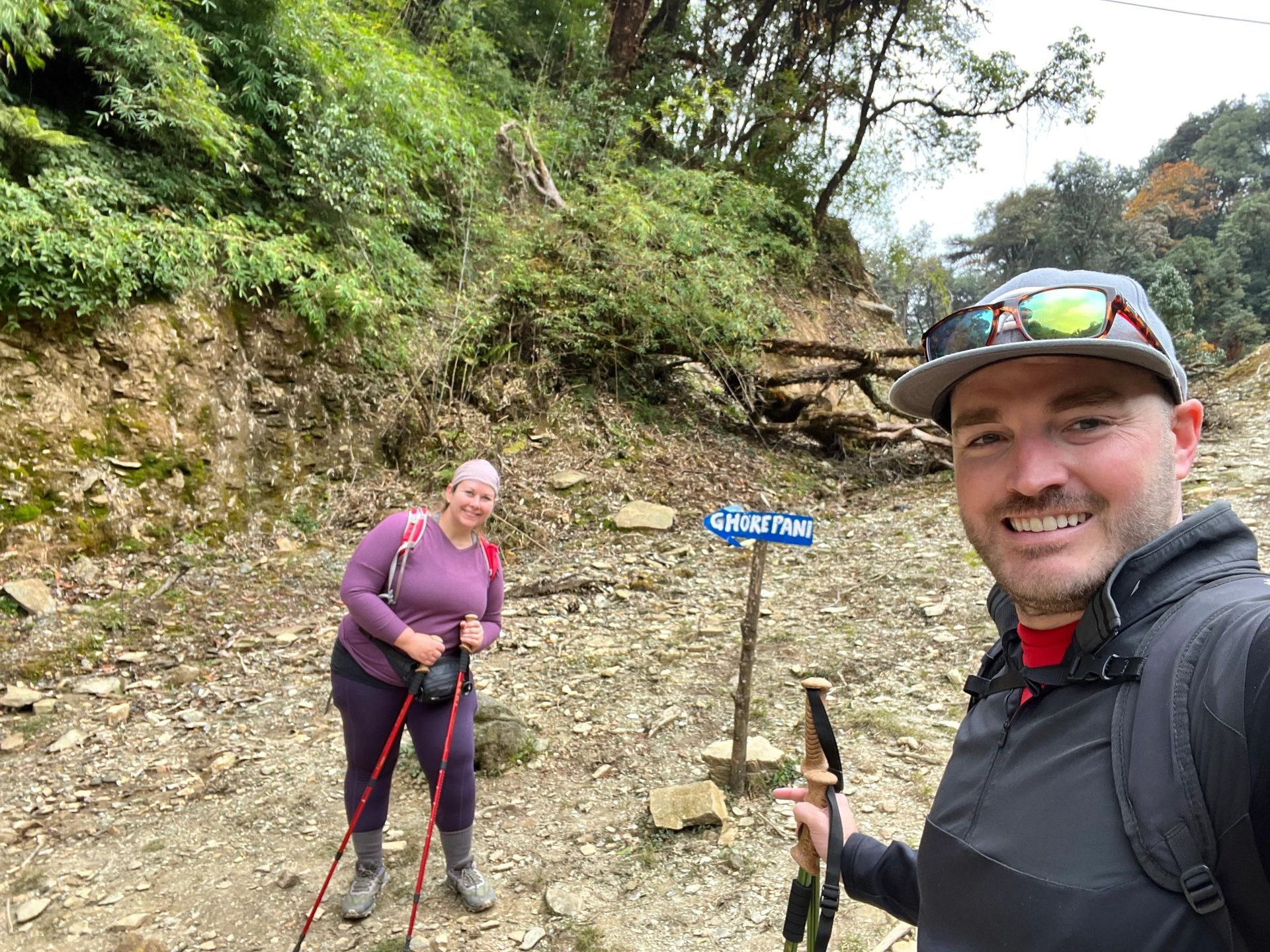Two hikers posing on a rocky trail in a wooded area, one holding trekking poles and smiling.