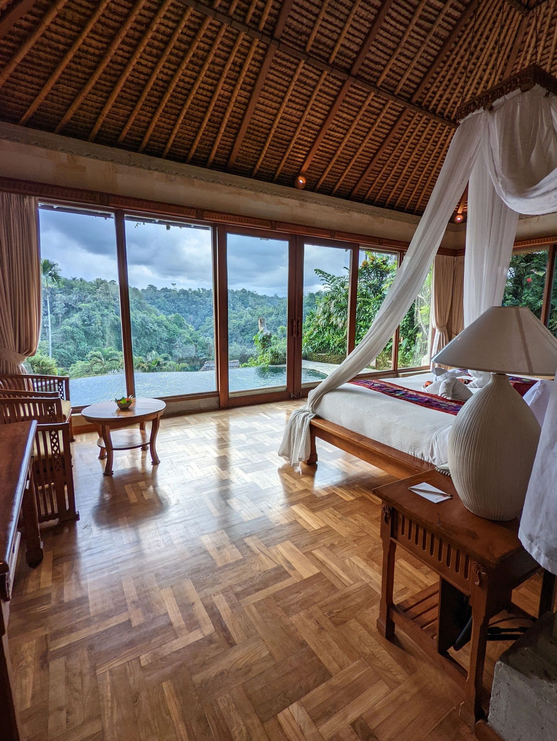 Rustic bedroom with wooden floor, canopy bed, and large windows overlooking lush tropical mountains and lake