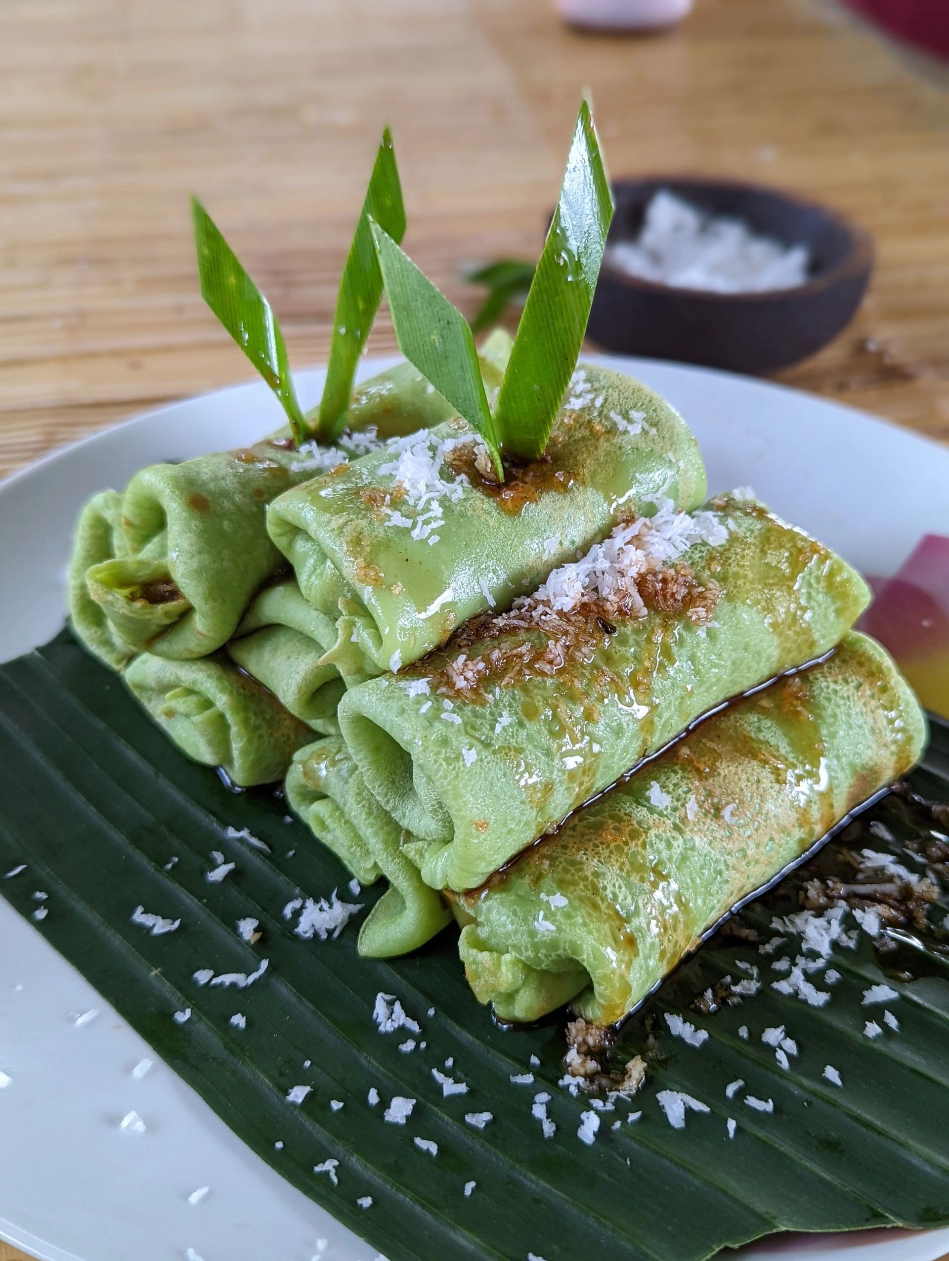 Green rolled crepes on a banana leaf, dusted with coconut, with pandan leaves garnish.