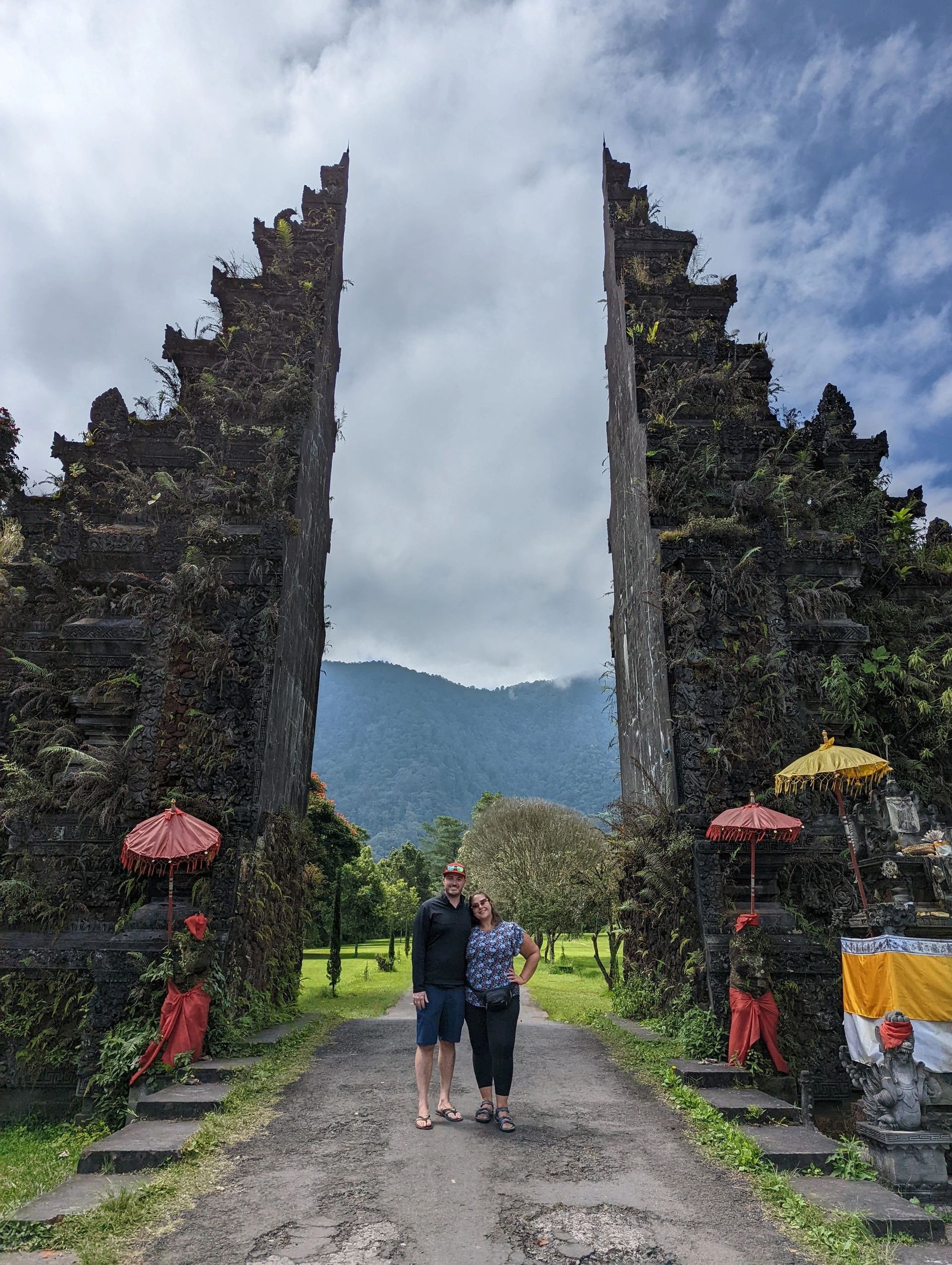 Two people walking through a stone temple gate with red umbrellas, mountains in the background