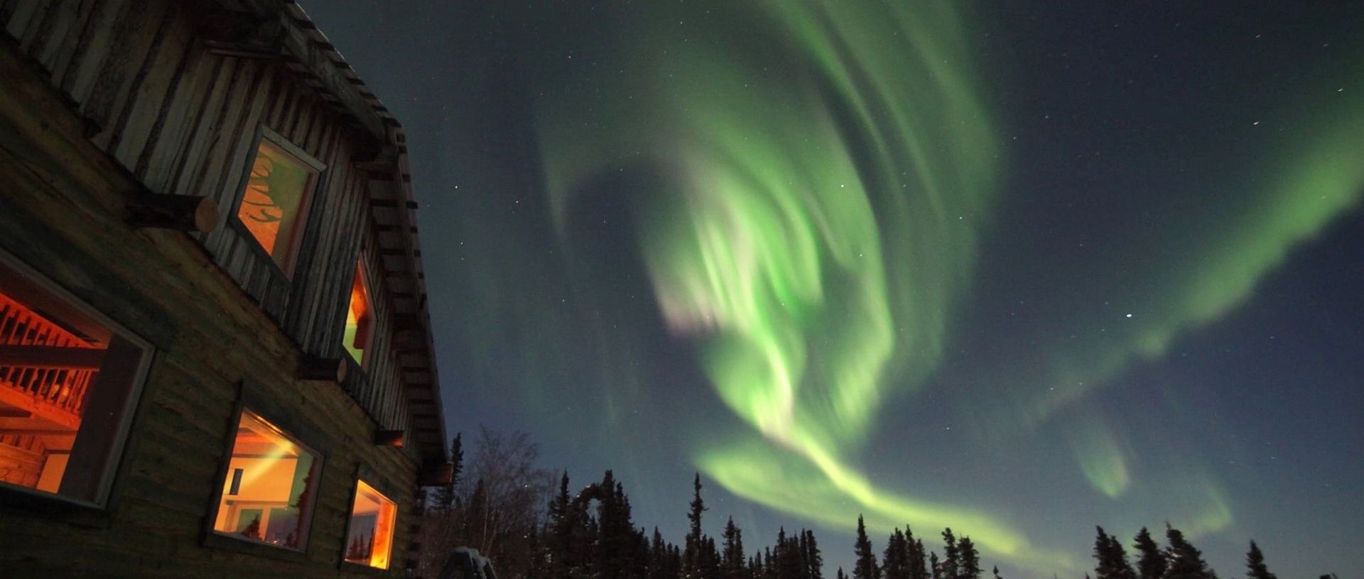 Northern lights swirling over a dark forest beside a warmly lit cabin at night