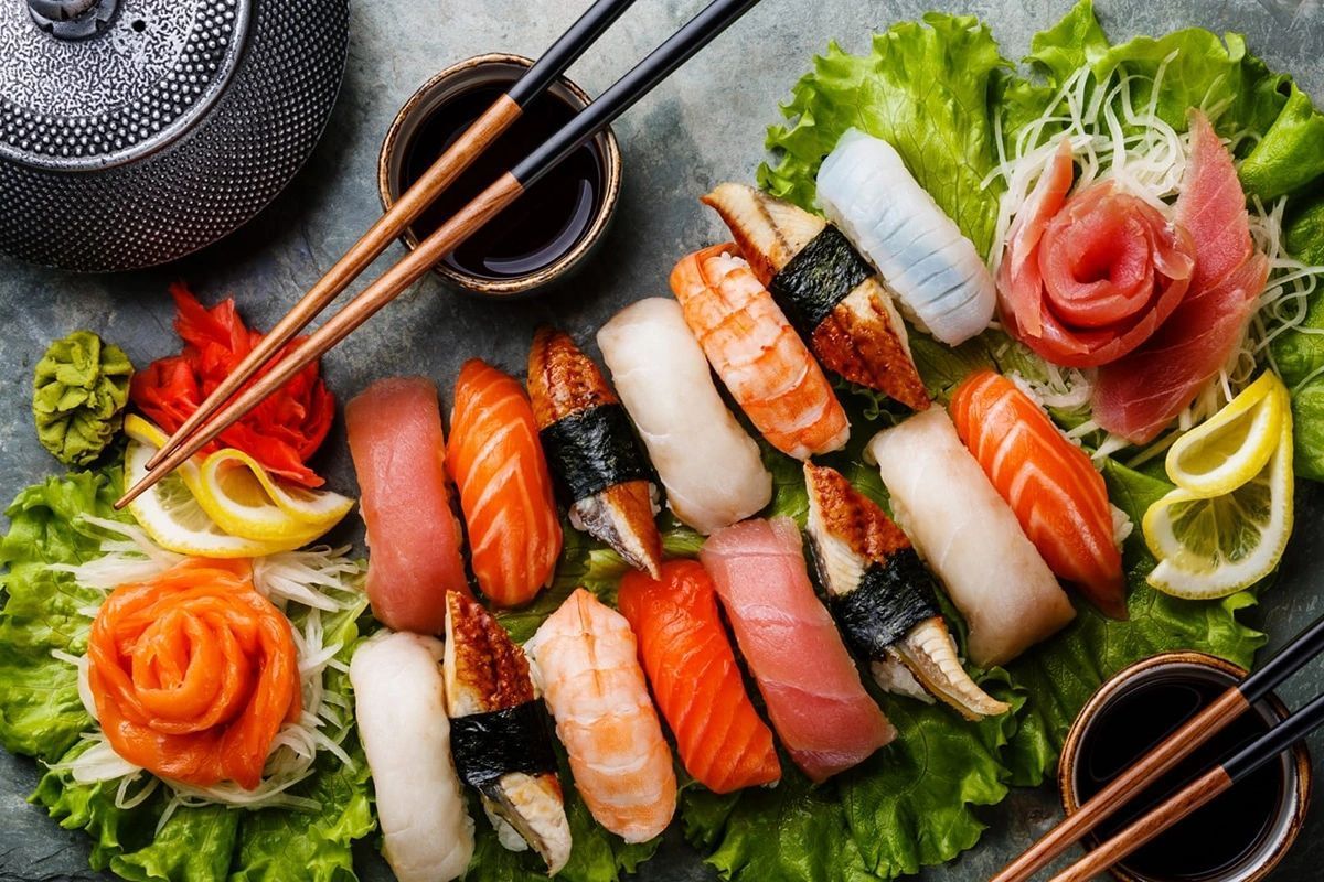 Assorted sushi rolls and sashimi on a black plate with chopsticks, lemon, and greens