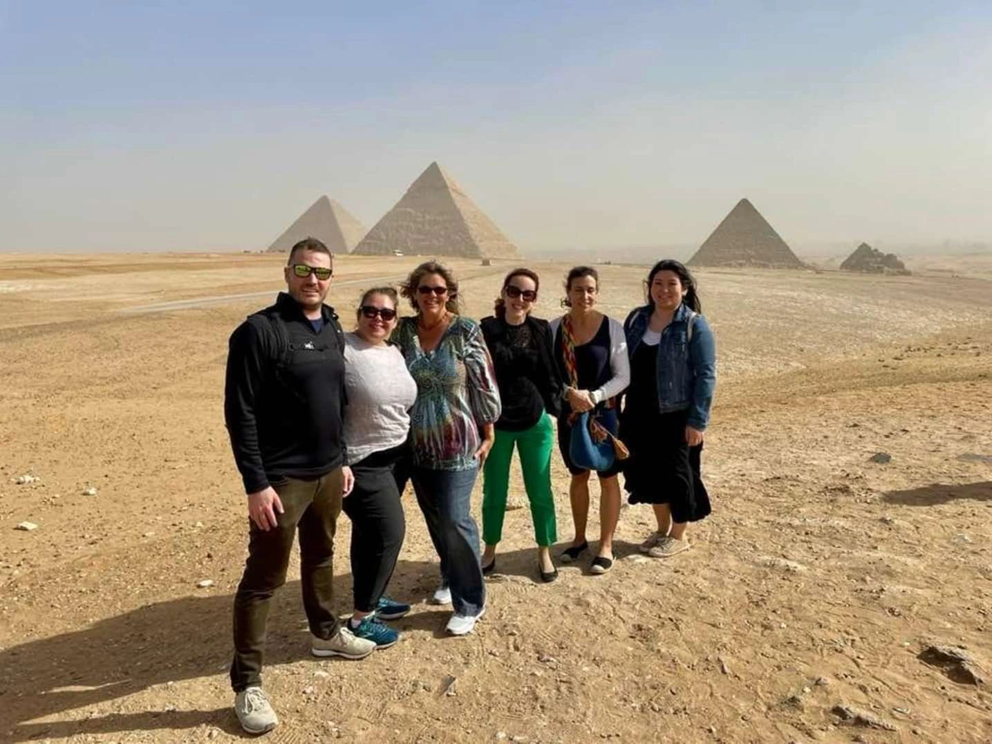 Six people pose in a desert with the pyramids visible in the background.