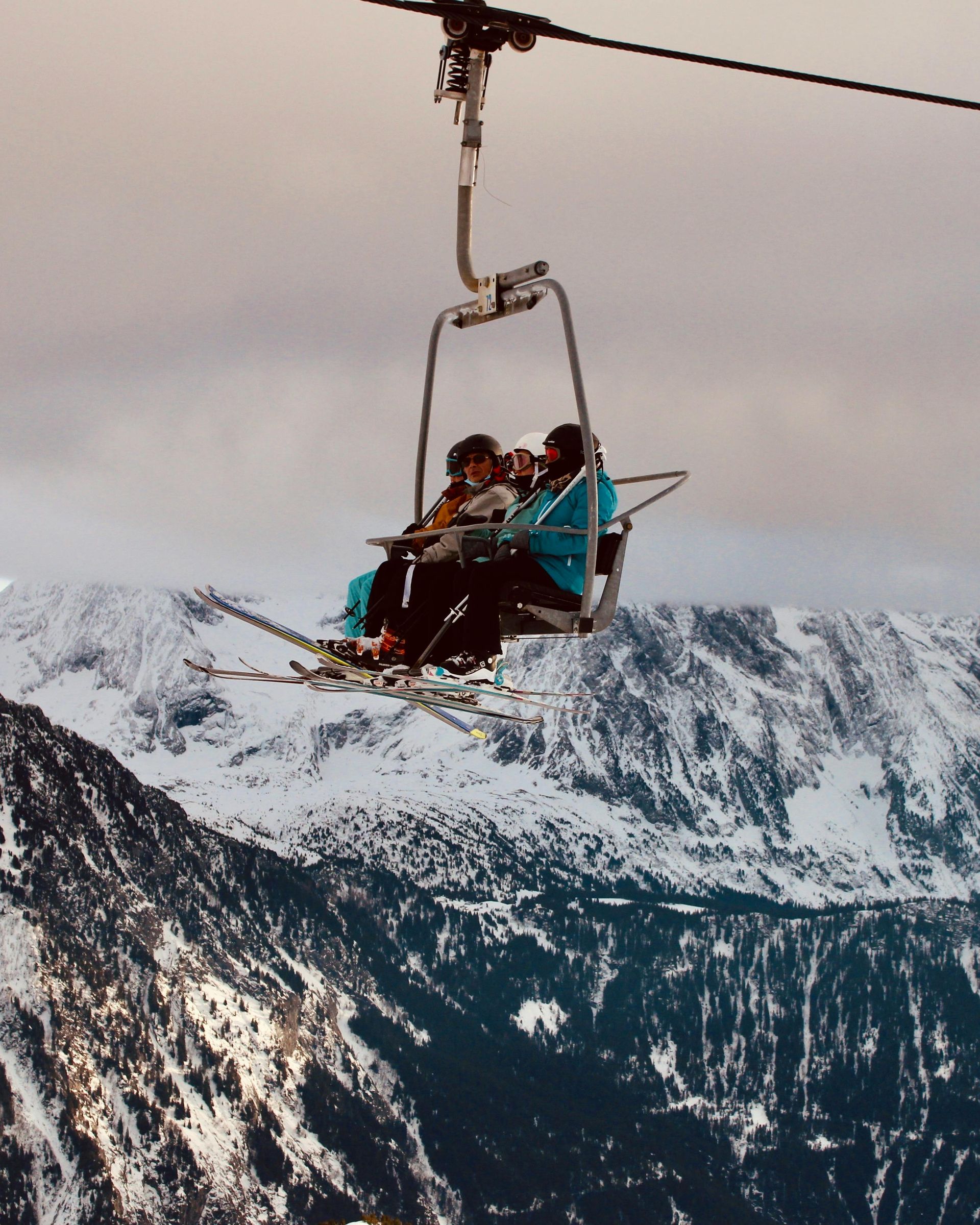 Two people ride a ski lift over snowy mountain slopes under a cloudy sky.