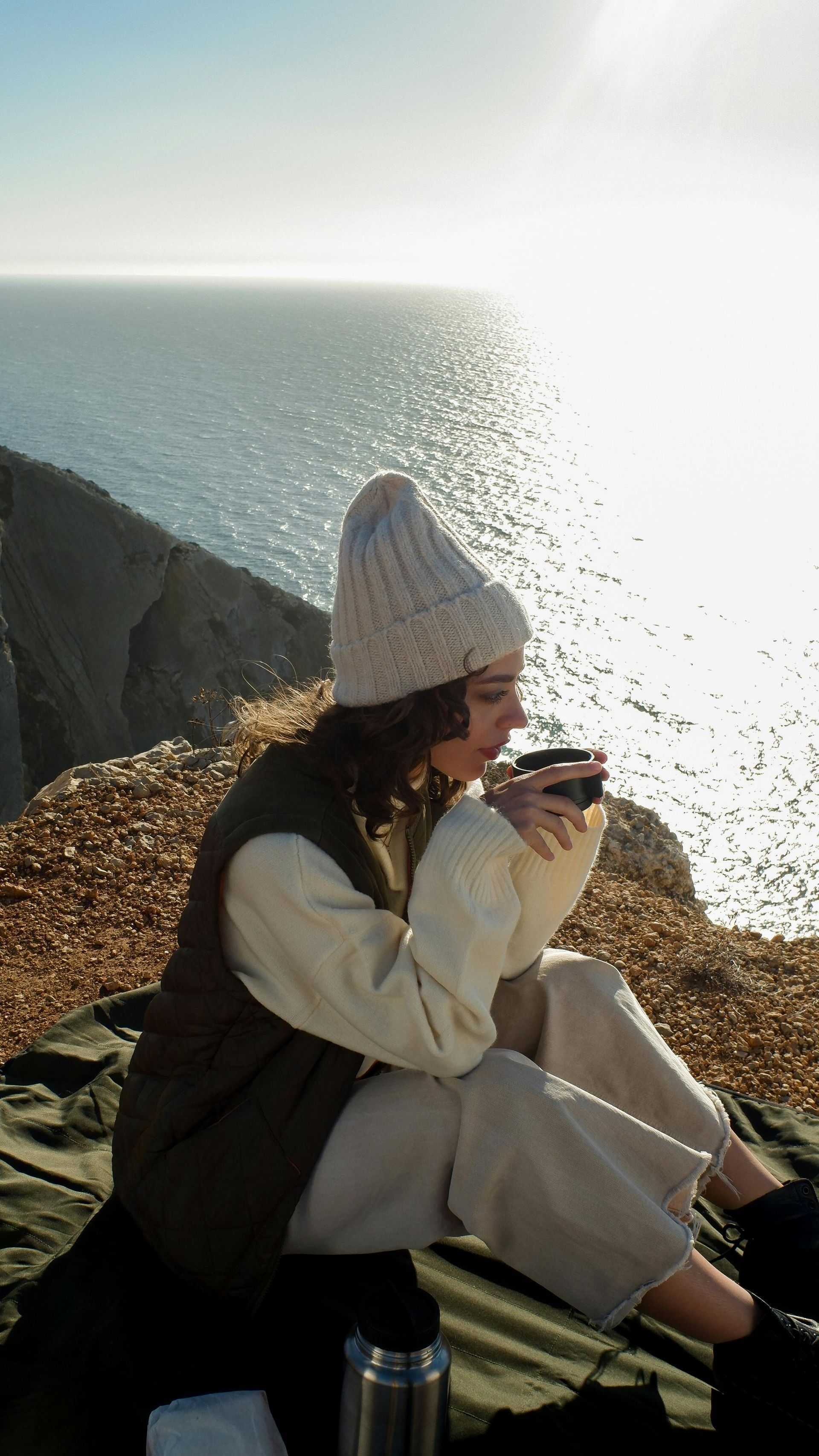 Person in a white knit hat sipping a drink beside a bright seaside cliff.