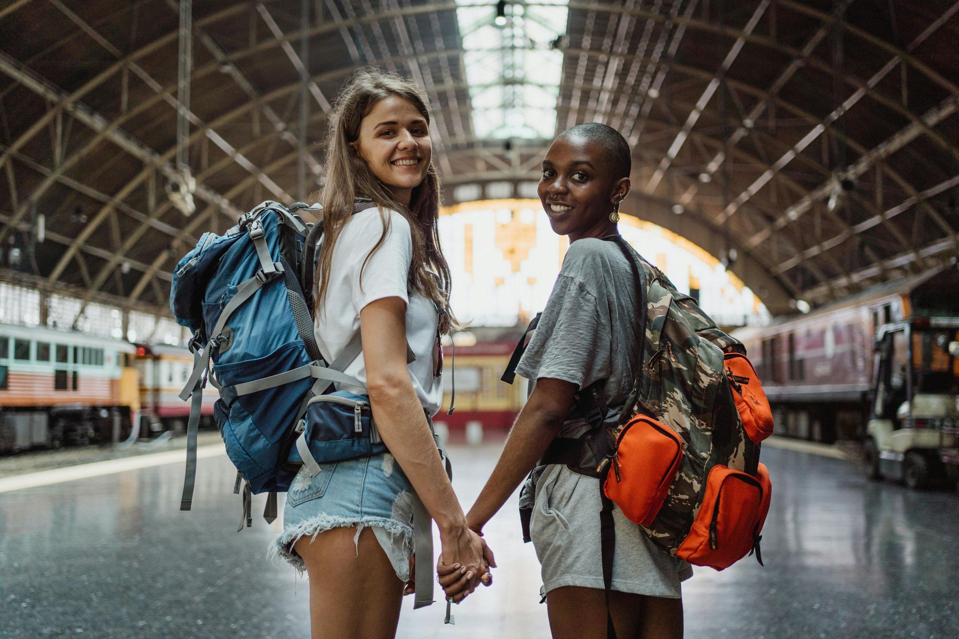 Two travelers with backpacks holding hands in a train station concourse