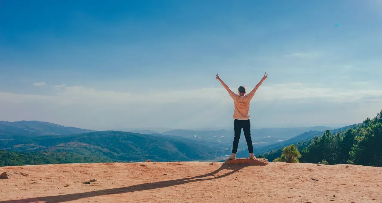 Person standing on a mountain overlook with arms raised, facing a wide blue sky and distant hills.