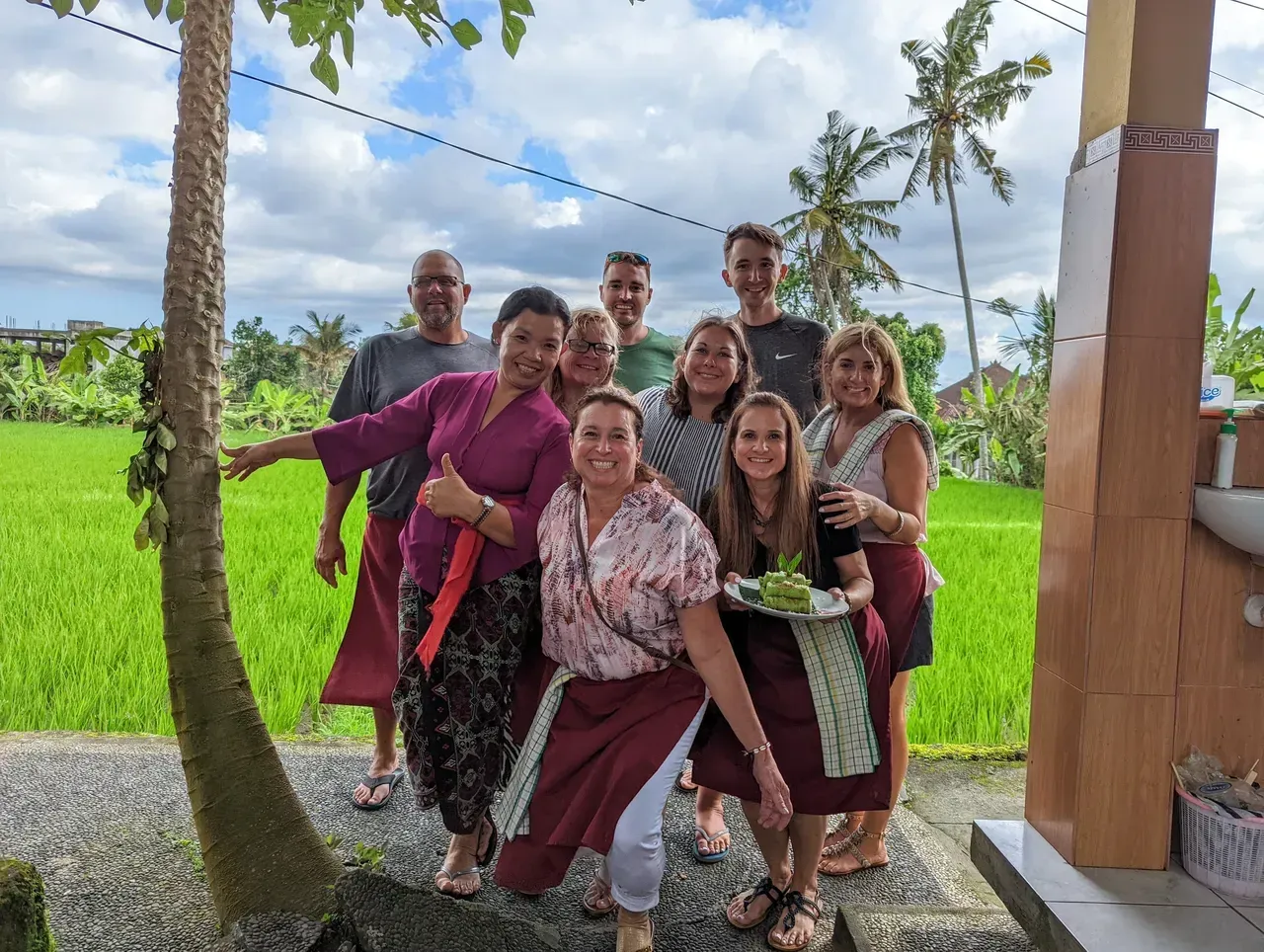 Group of smiling people posing outdoors beside green fields and palm trees under a bright sky