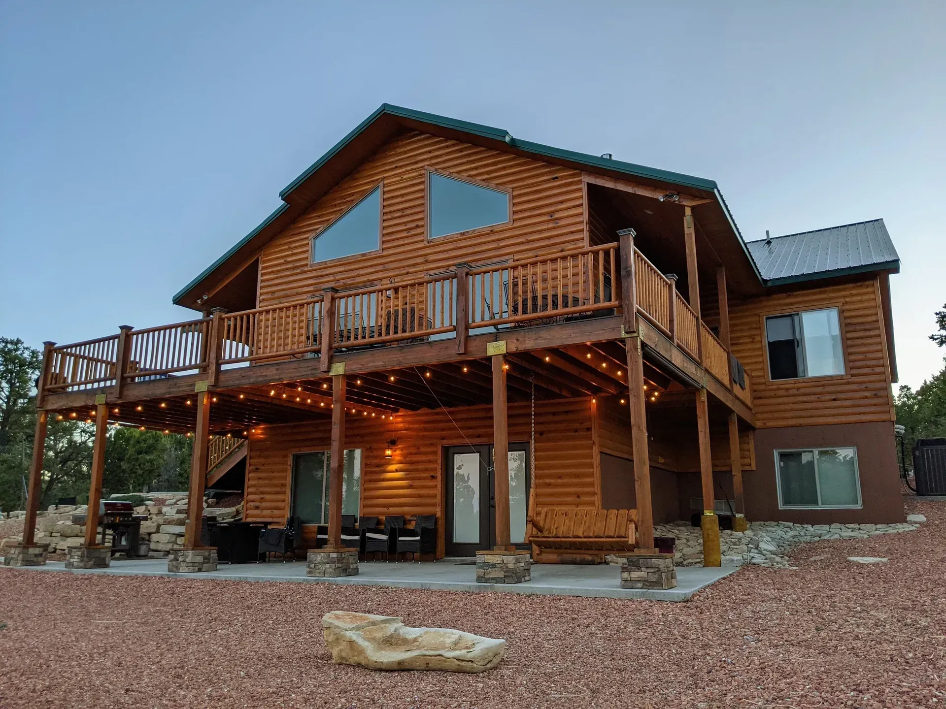 Wooden cabin-style house with a wraparound deck and lit porch at dusk
