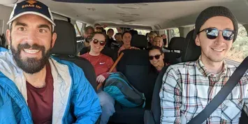 Group of friends smiling in a van, with a bearded man in front and passengers in back seats