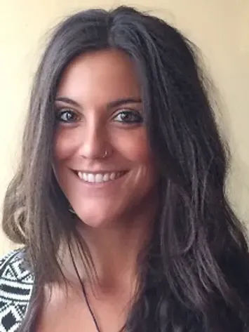 Smiling person with long dark hair and a nose ring, wearing a patterned top, against a light background
